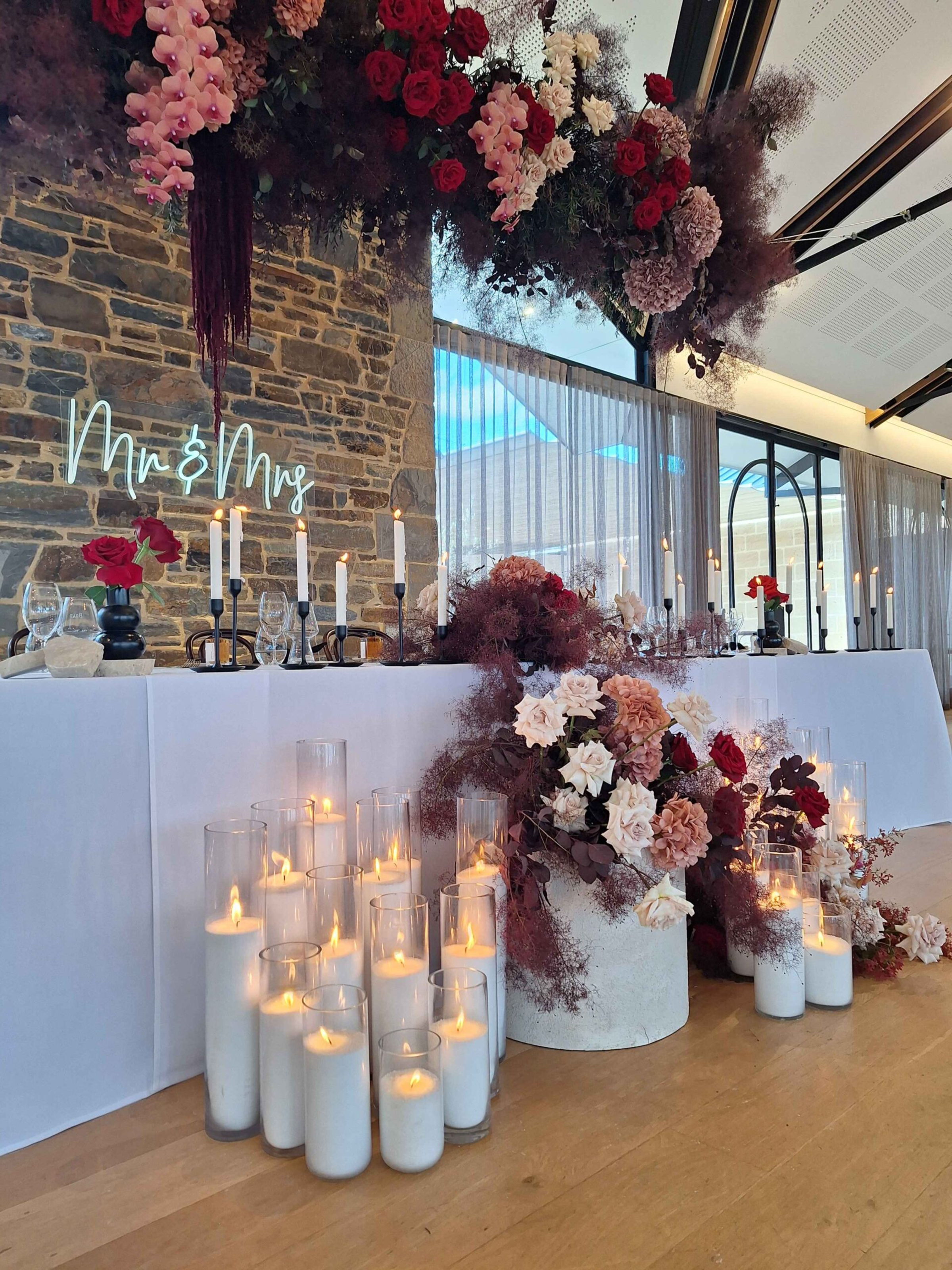 Romantic wedding sweetheart table with candles, floral arrangements, and a neon Mr & Mrs sign against a stone wall backdrop.
