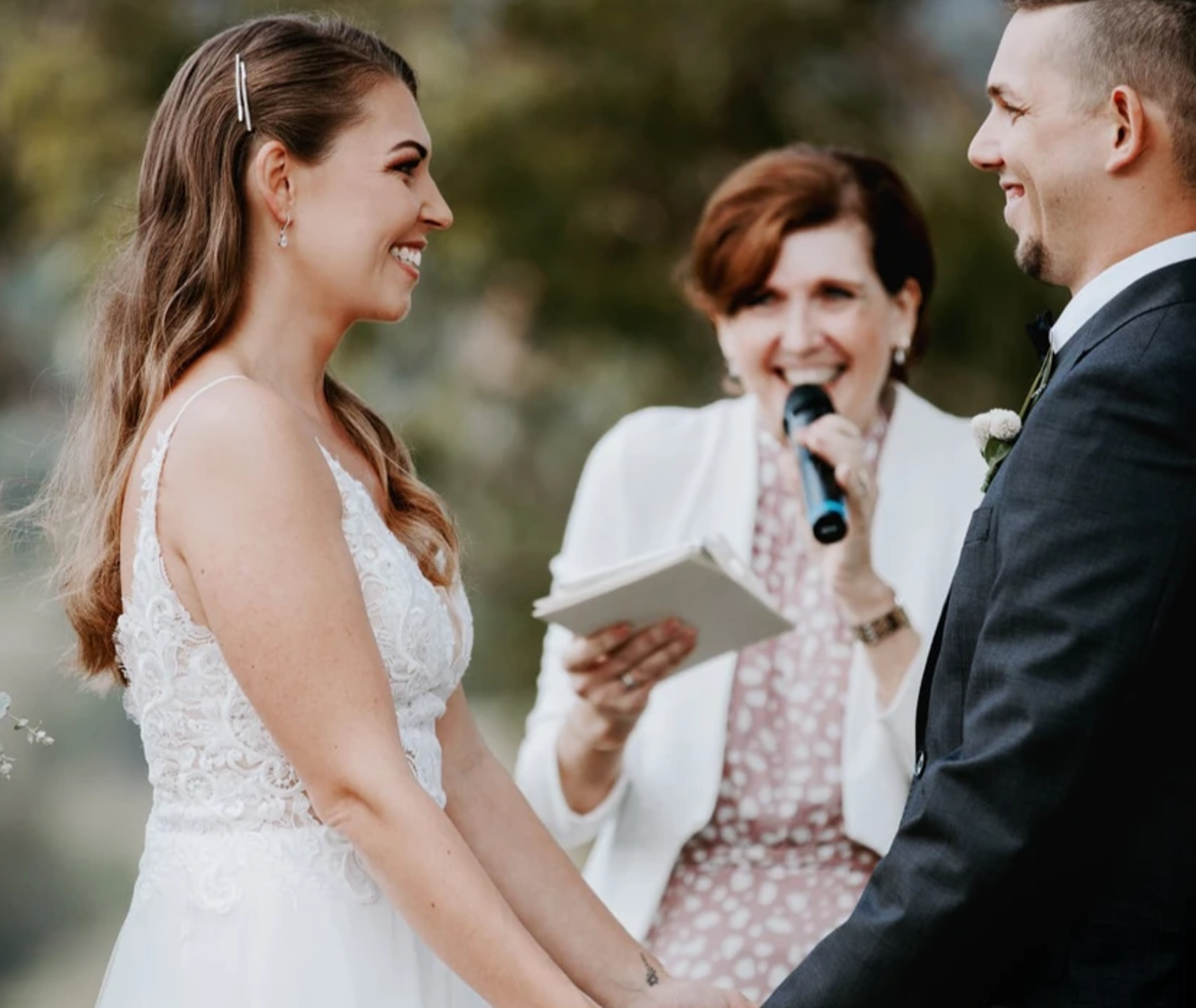 Bride and groom holding hands during an outdoor ceremony with an officiant speaking into a microphone.