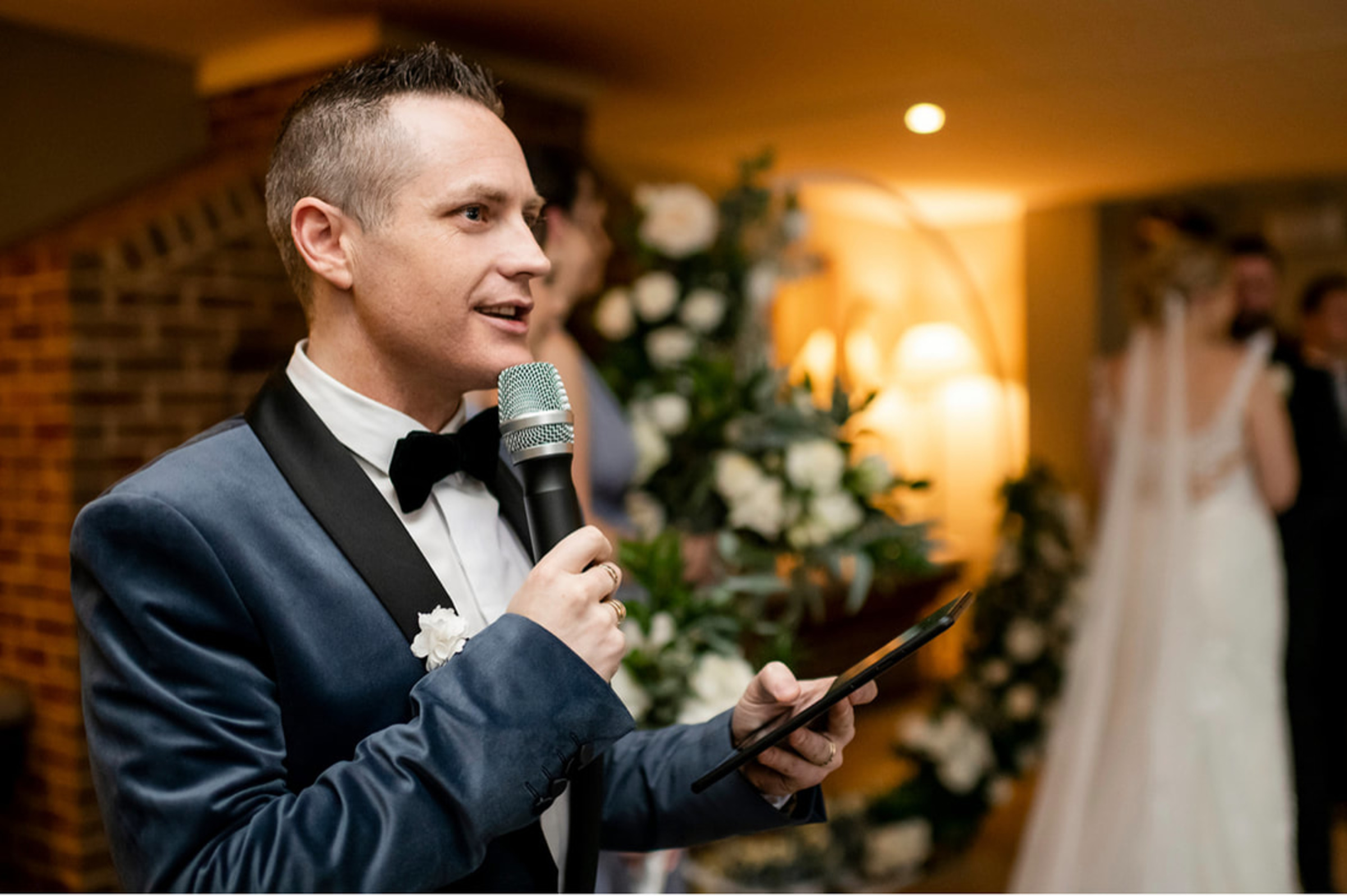 Wedding MC in a tuxedo speaking into a microphone at an indoor reception with floral decor and bride in the background.