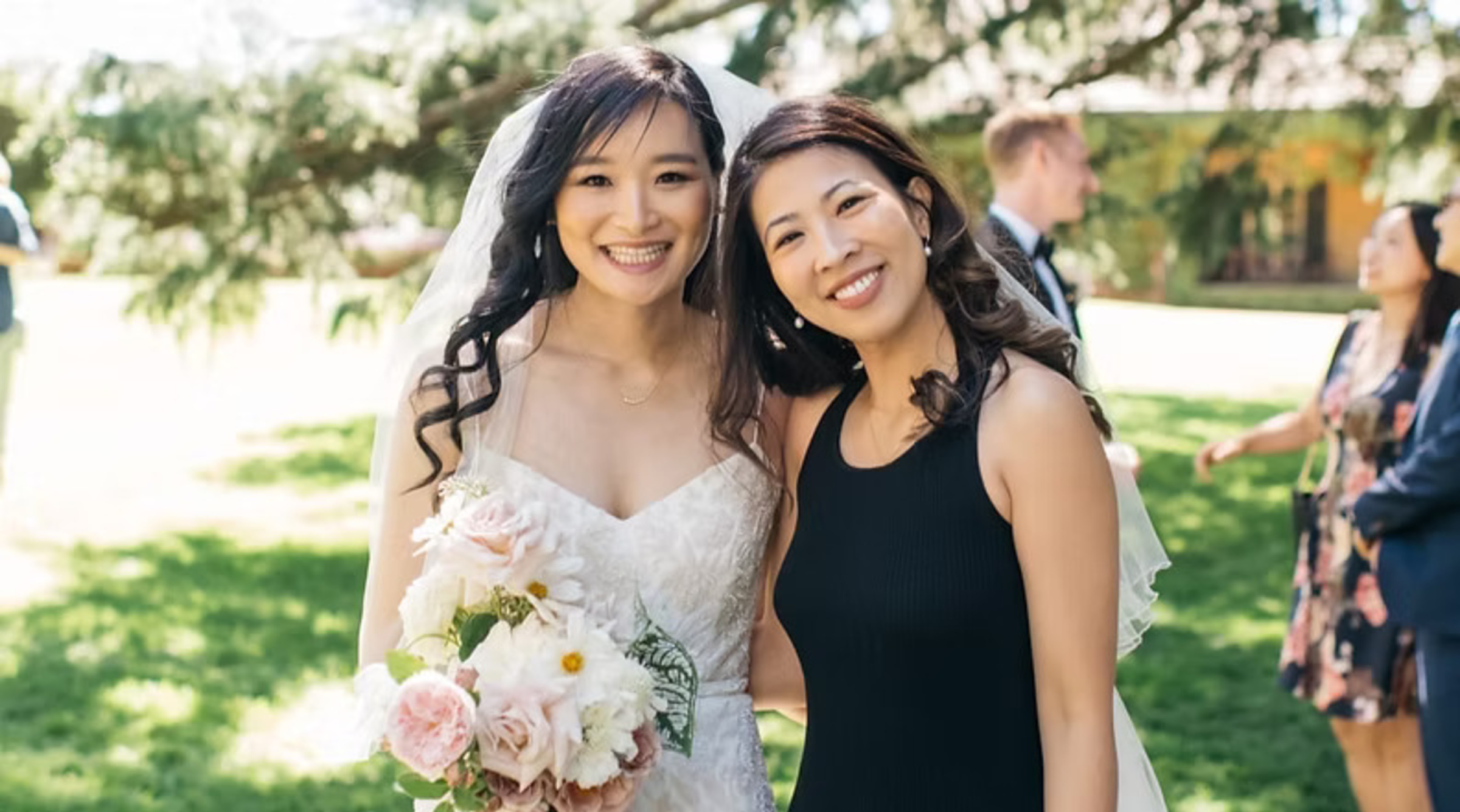 Bride holding a pastel bouquet smiles with a friend at an outdoor garden wedding.