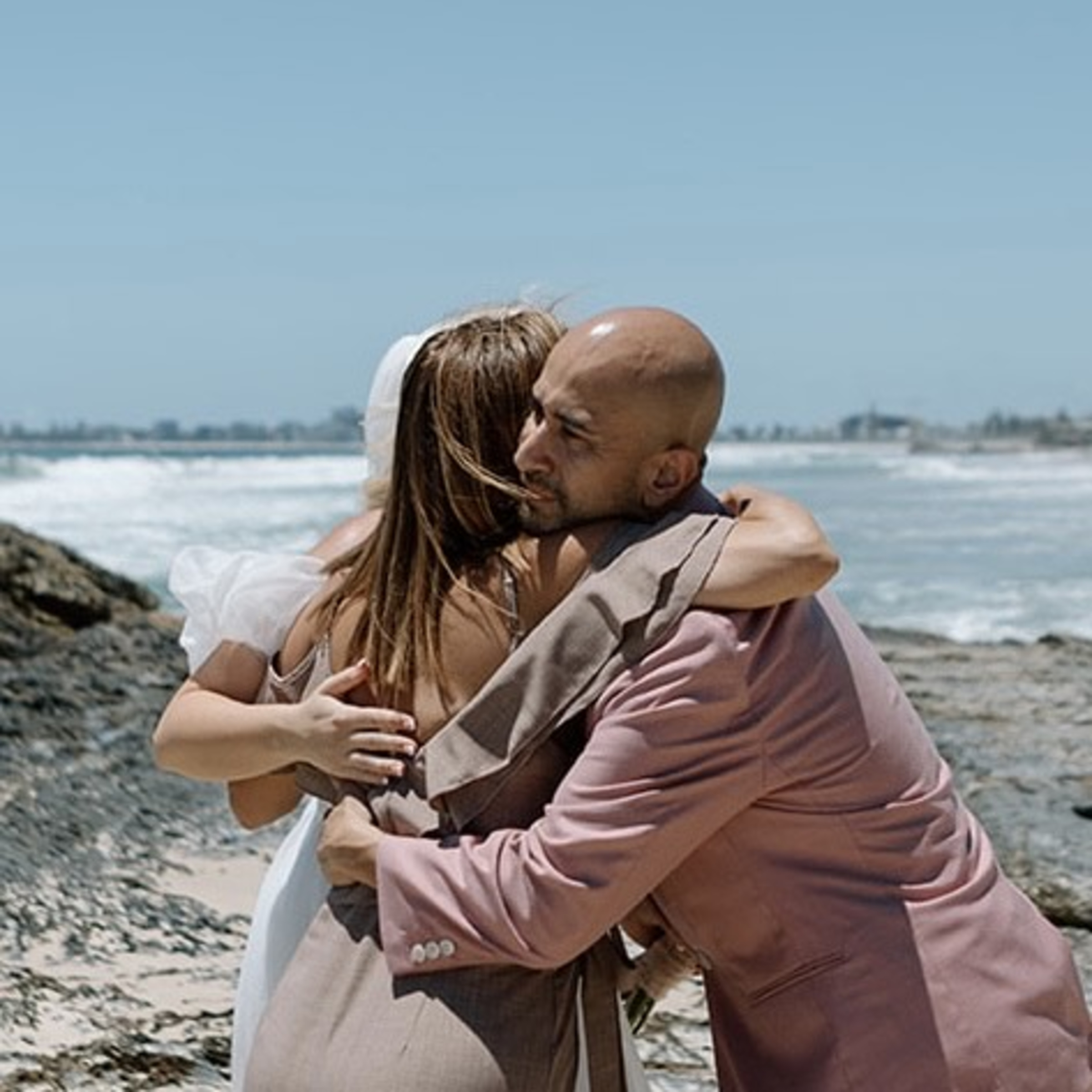 Wedding guests embrace in a heartfelt hug on a rocky beach with waves and blue sky in the background.