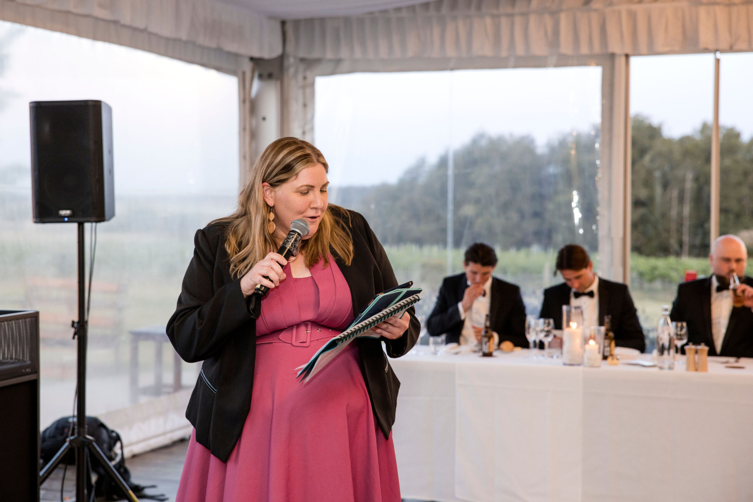 A woman delivers a wedding speech with a microphone in front of a seated head table at a reception.