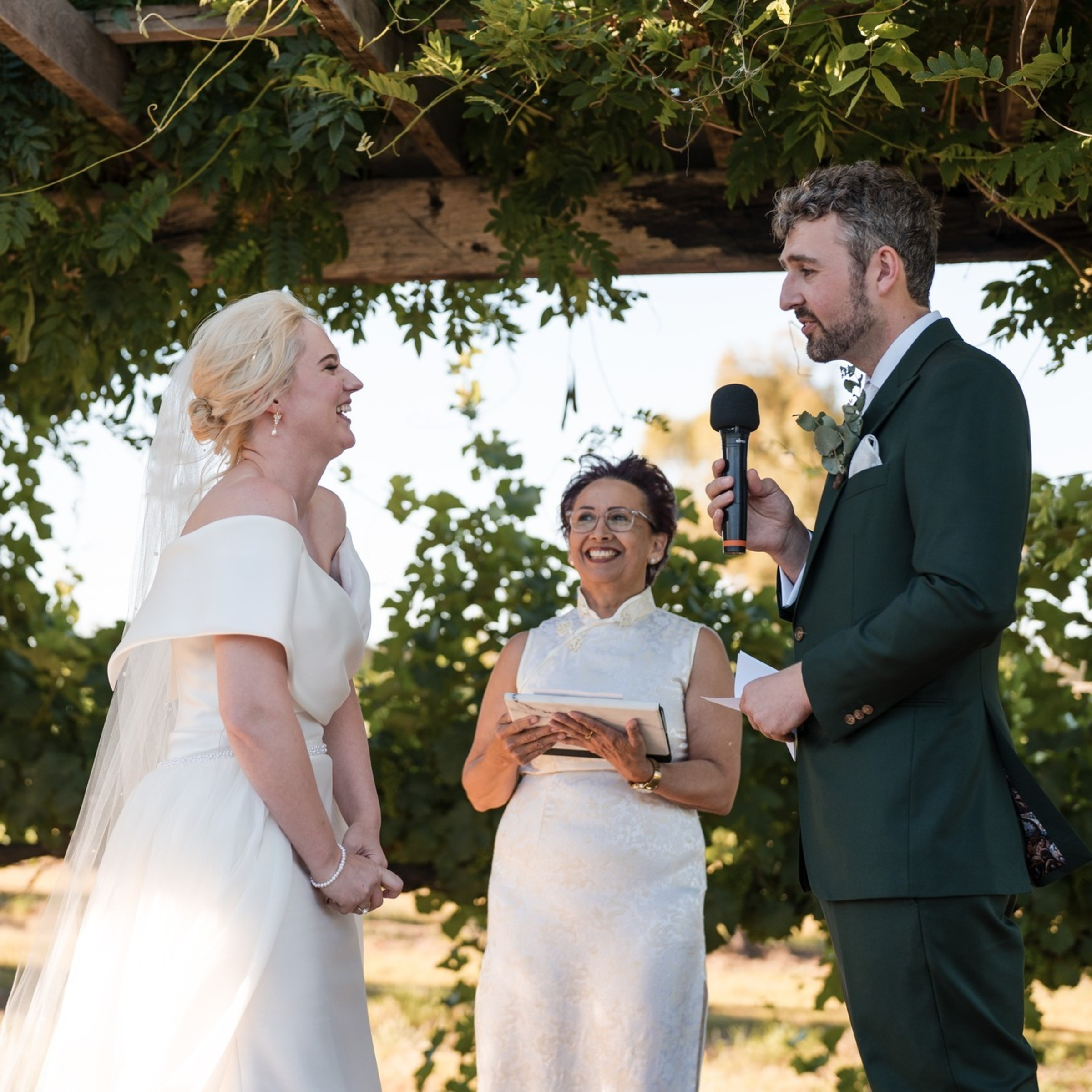 Bride and groom exchange vows with an officiant under a leafy outdoor arbor.