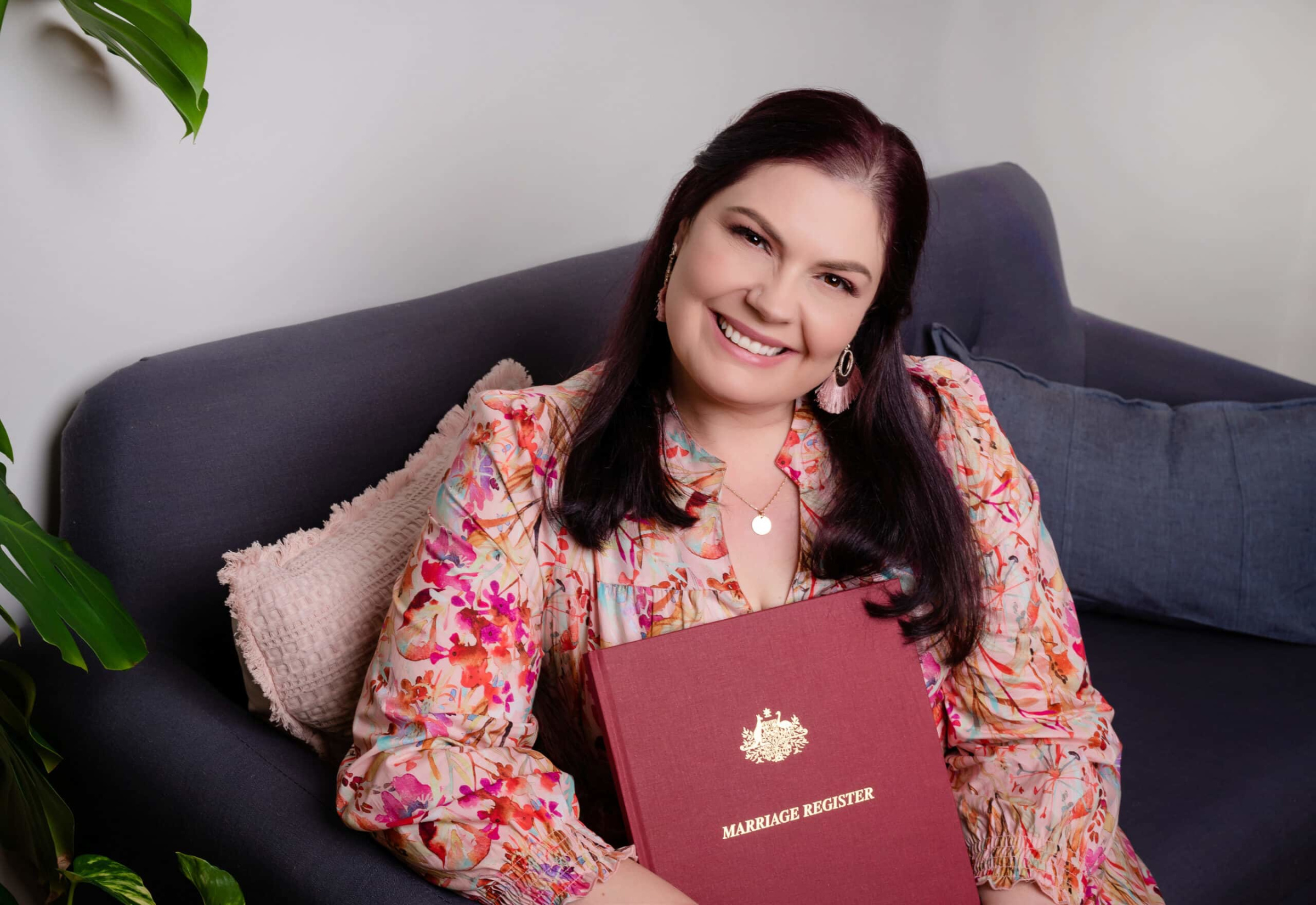 Smiling wedding celebrant sitting on a sofa holding a red marriage register book.