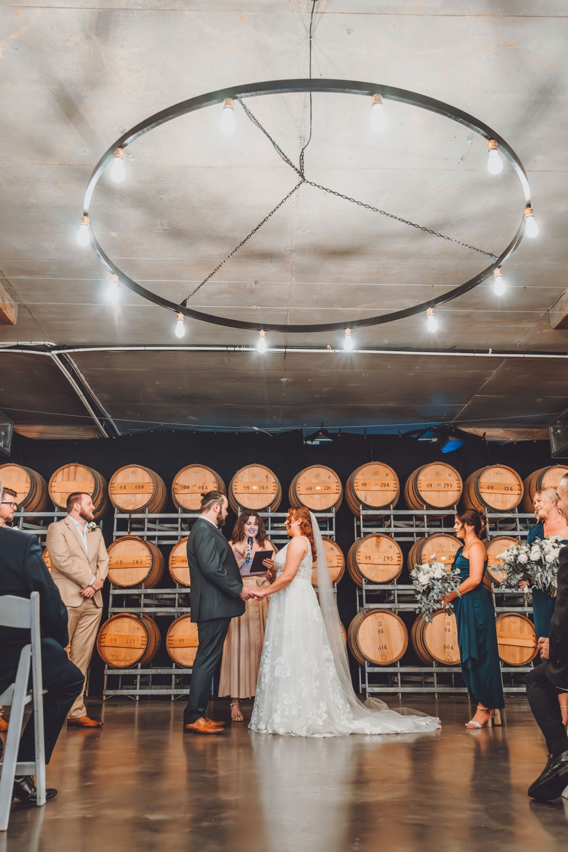 A couple exchanges vows in a winery barrel room surrounded by their wedding party and warm string lights.