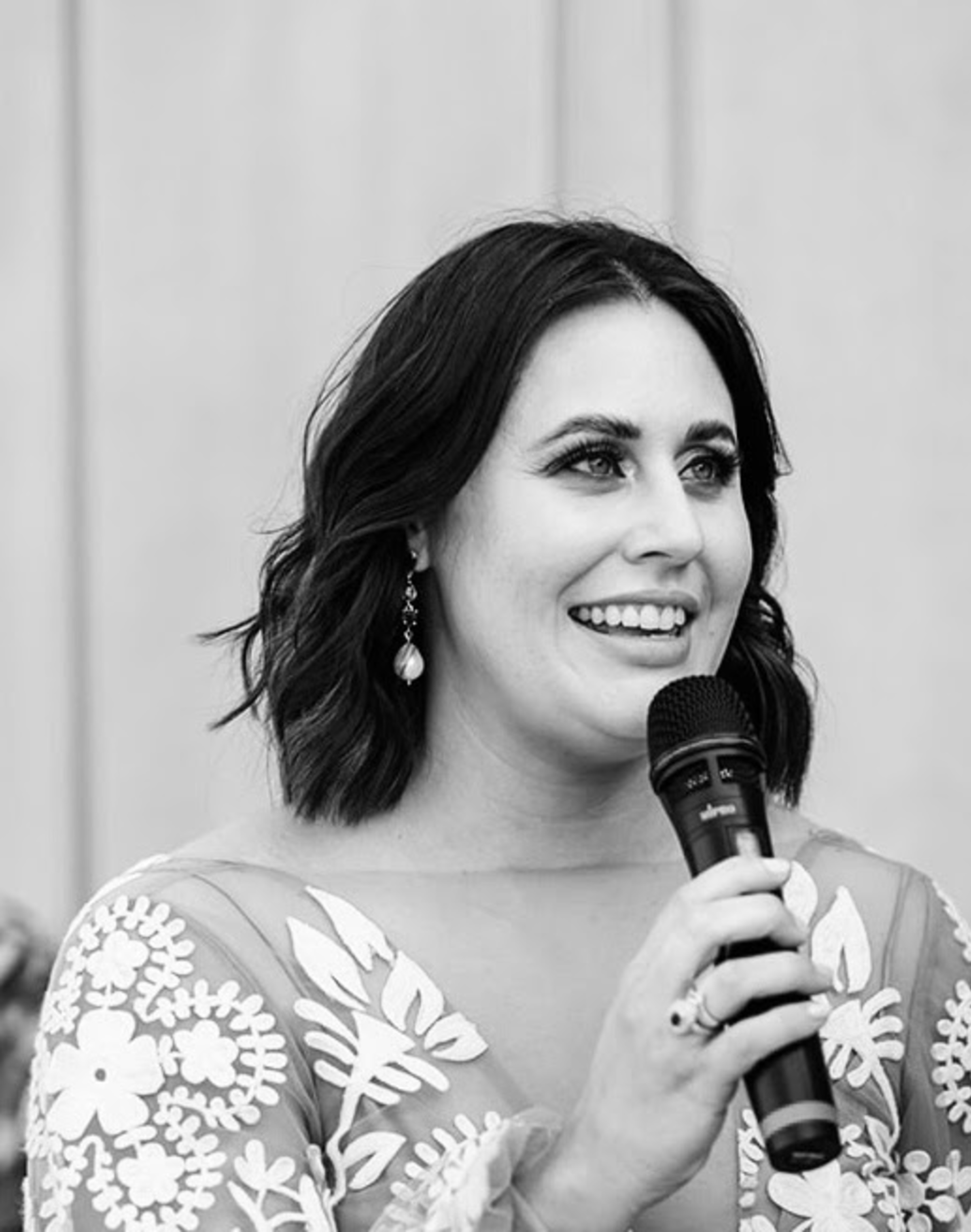 Smiling woman holding a microphone while speaking at a wedding ceremony.