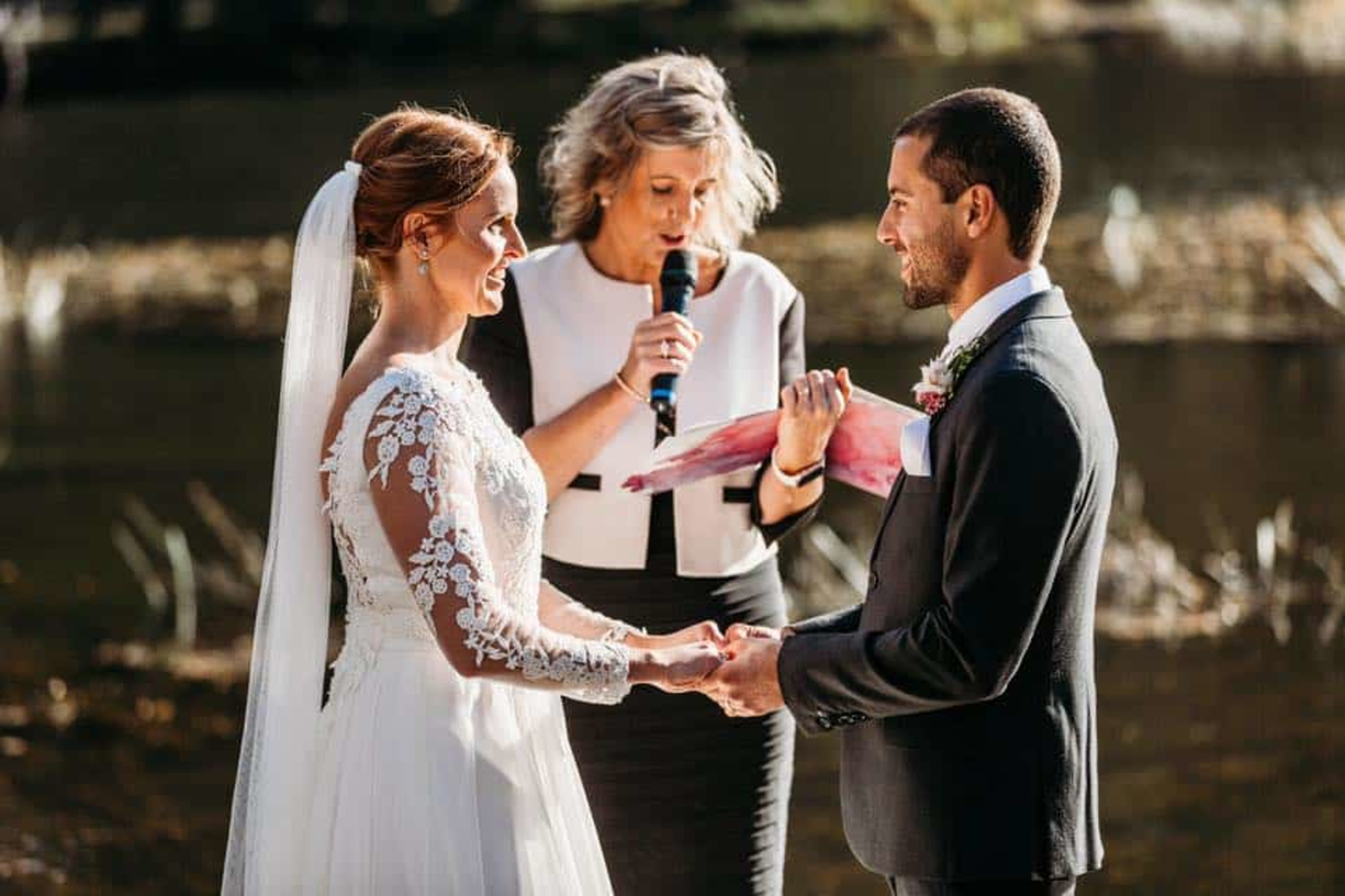 Bride and groom hold hands during an outdoor lakeside ceremony as the officiant reads their vows.