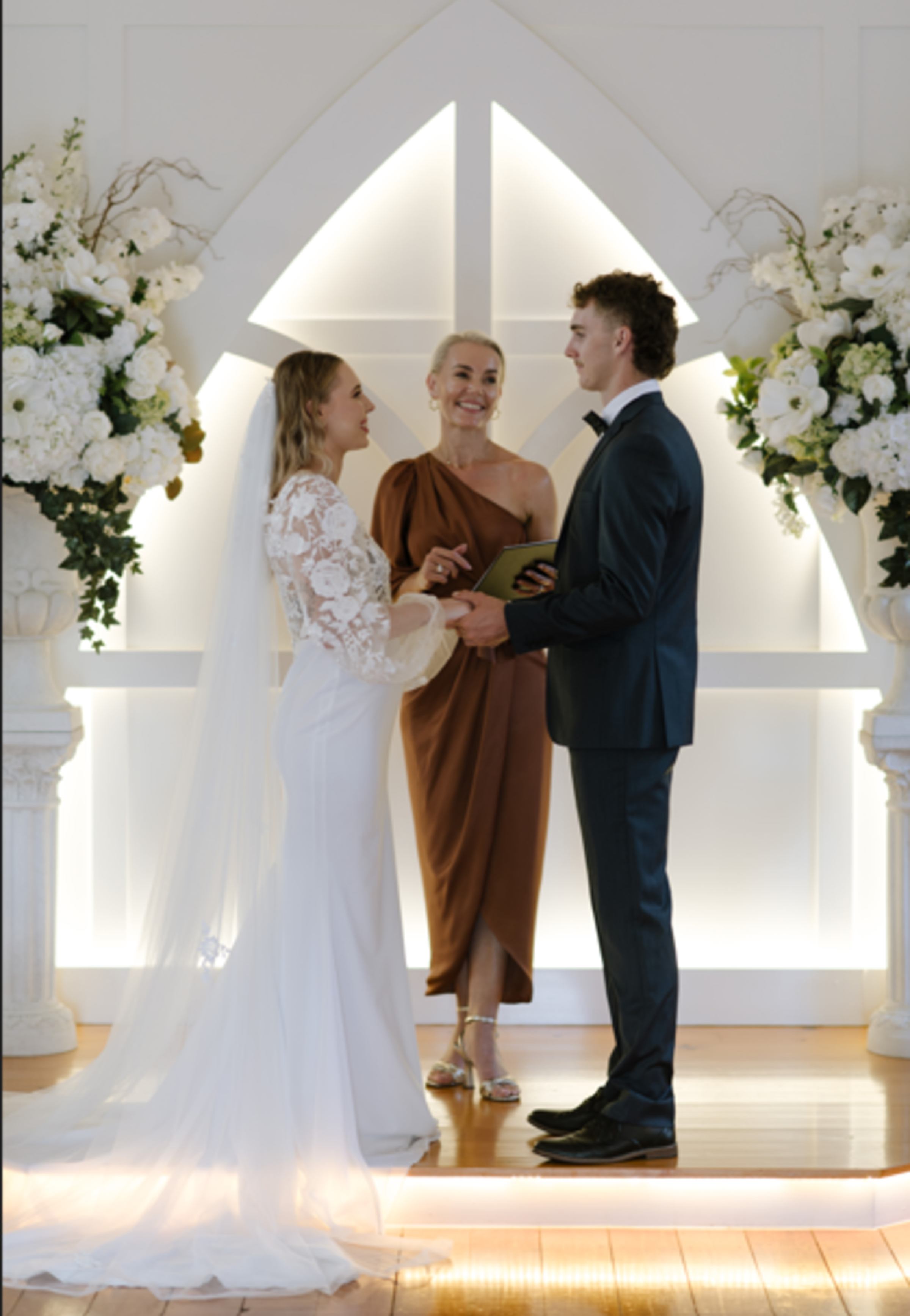 Bride and groom stand with an officiant in a modern indoor chapel framed by large white floral arrangements.