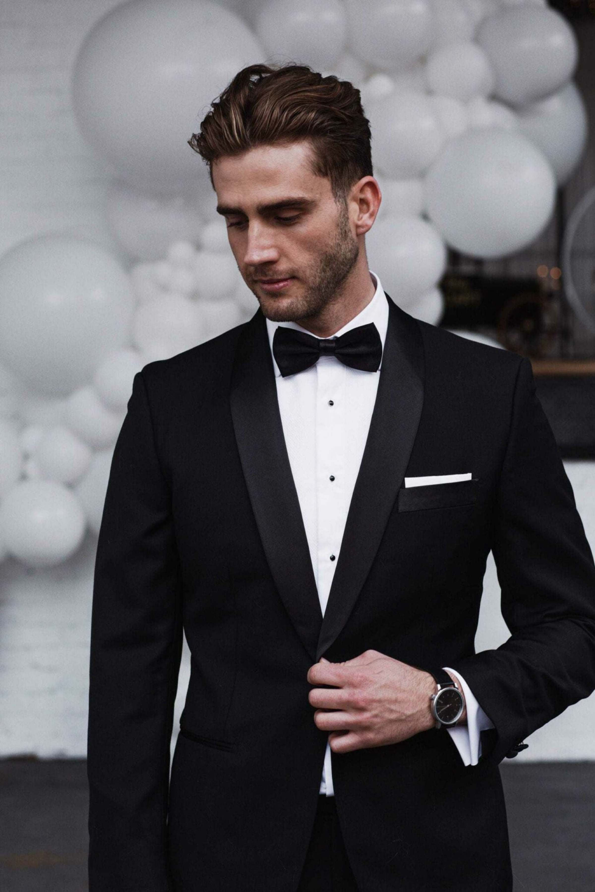 Groom in a classic black tuxedo with bow tie standing before white balloon backdrop.