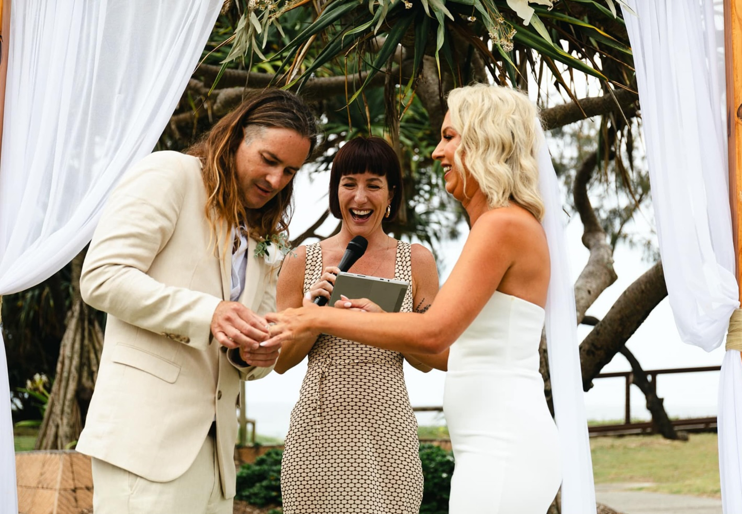 Couple exchanges rings during an outdoor ceremony with a smiling celebrant under a simple draped arbor.