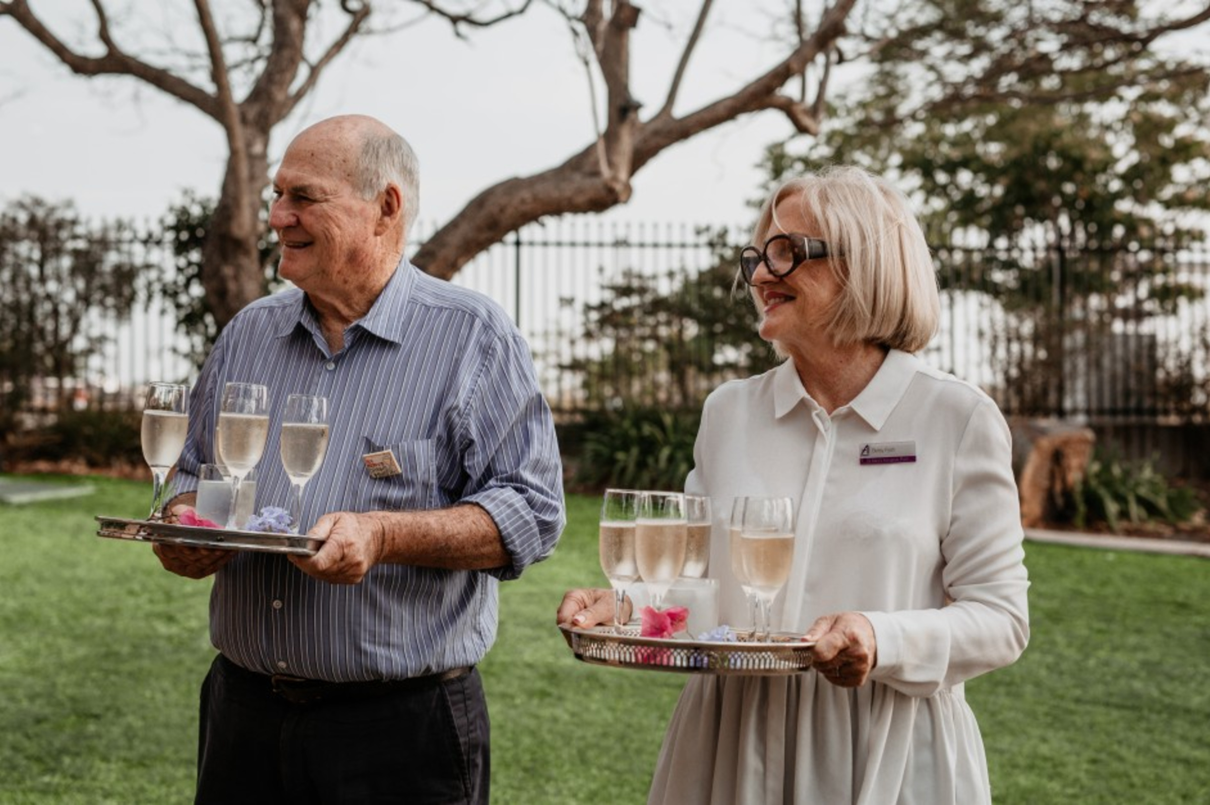 Two smiling servers carry trays of champagne glasses at an outdoor wedding reception.