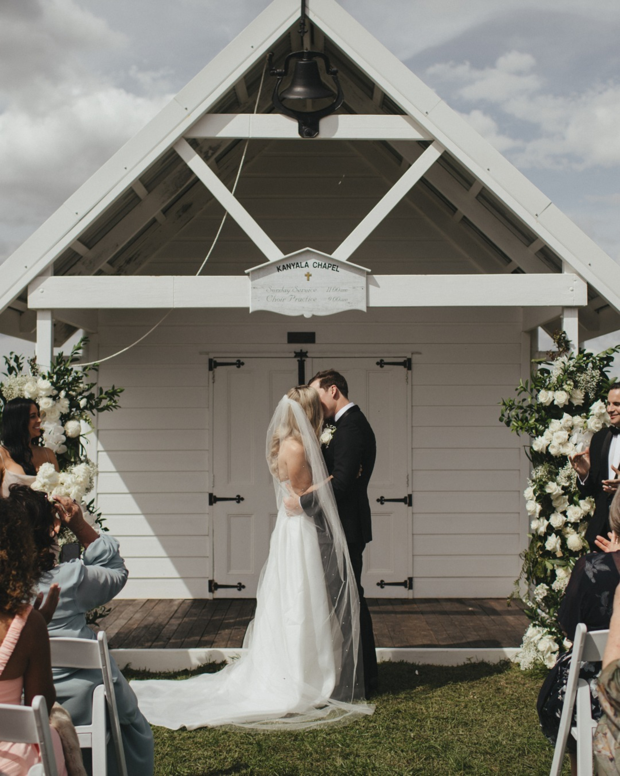 Bride and groom share a kiss in front of a white chapel during their outdoor wedding ceremony.