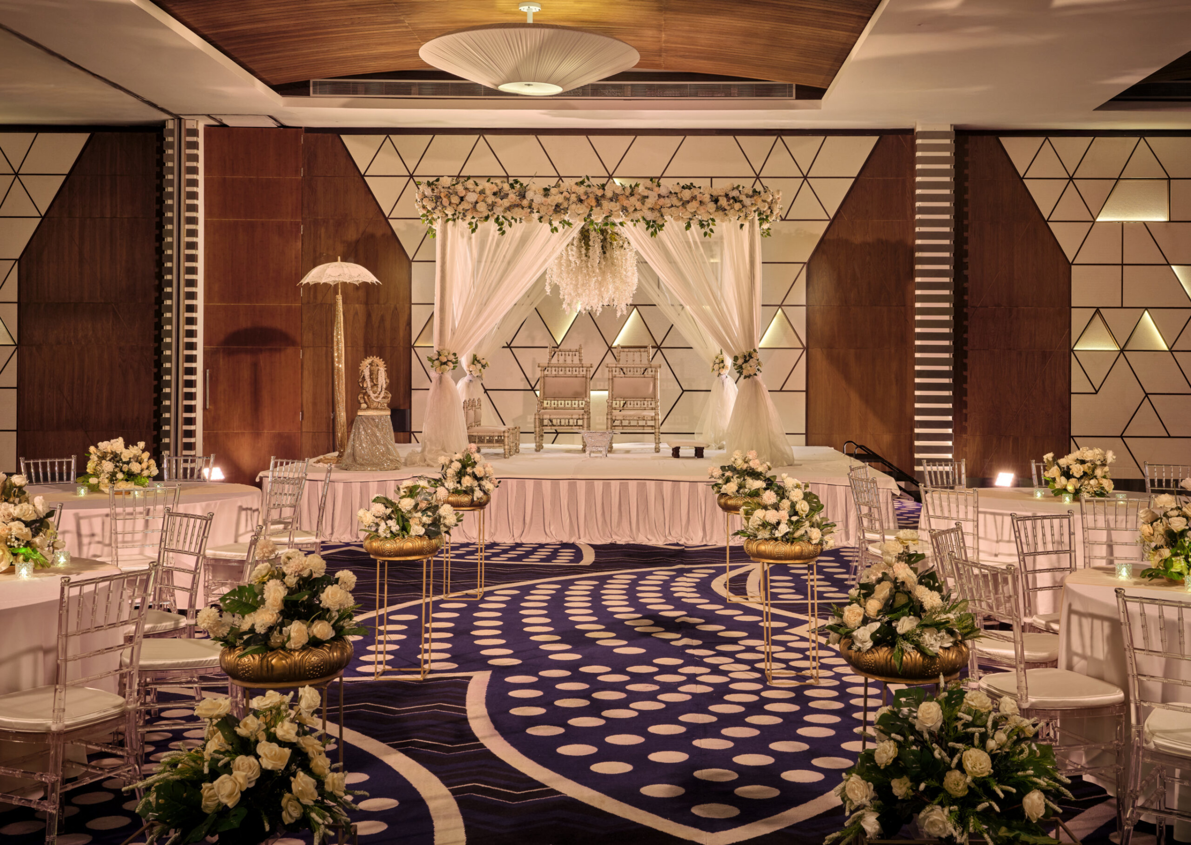 Luxurious indoor wedding ceremony setup with floral mandap, elegant chairs, and modern banquet hall decor.