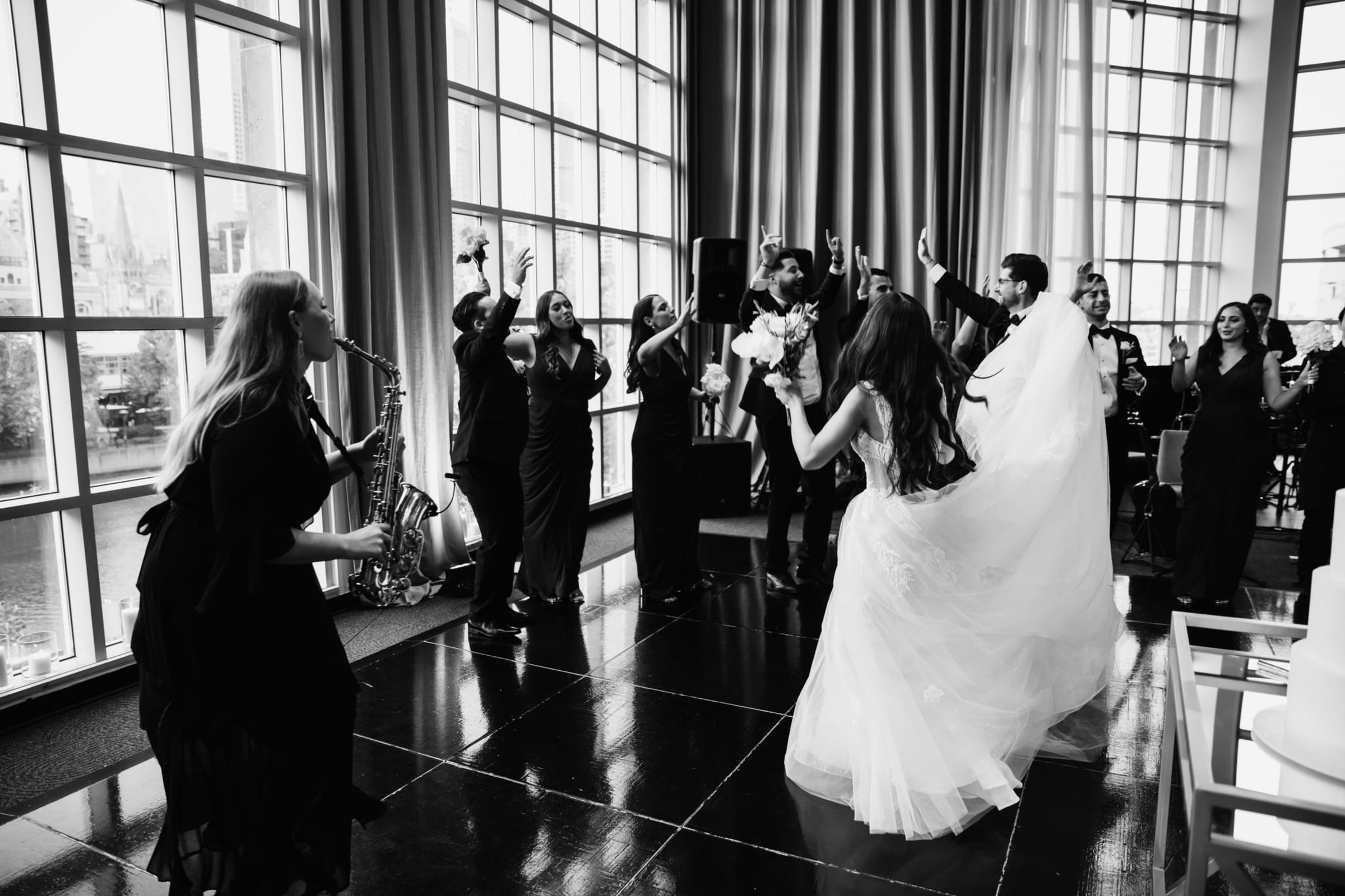 Black and white photo of a bride and guests dancing to a live saxophonist at an indoor wedding reception.