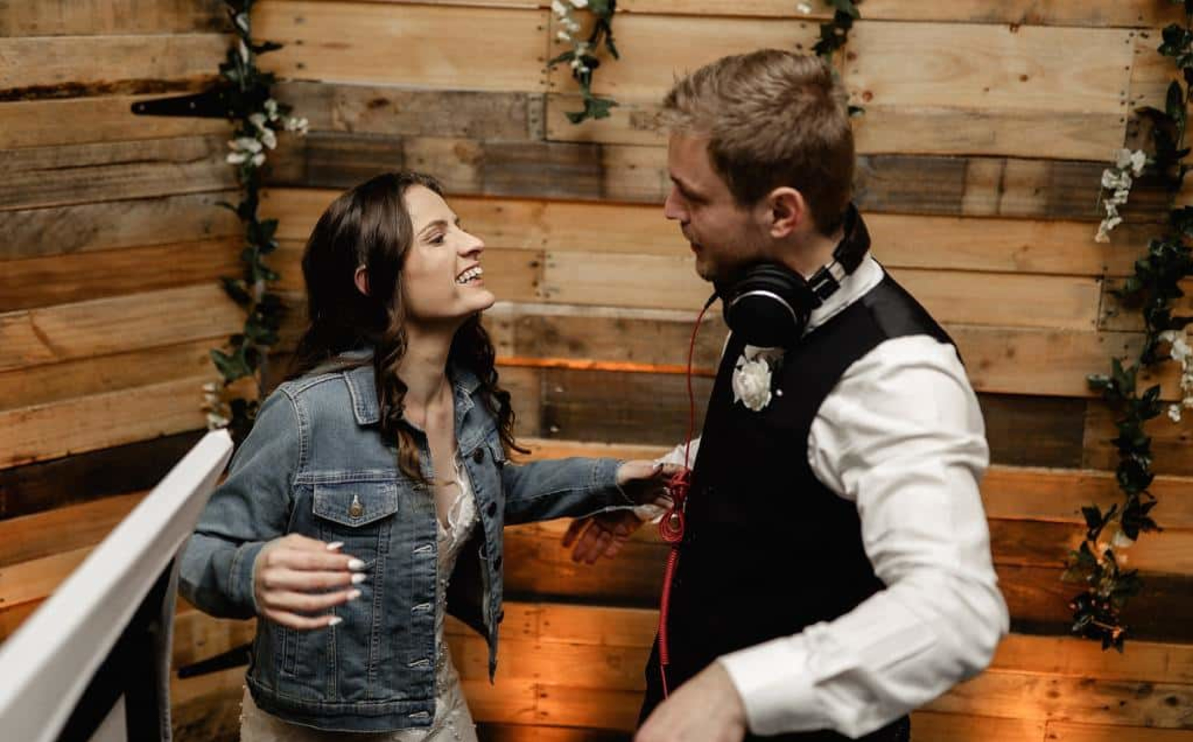 Bride in denim jacket laughs and dances with a DJ in a rustic wooden wedding reception setting.