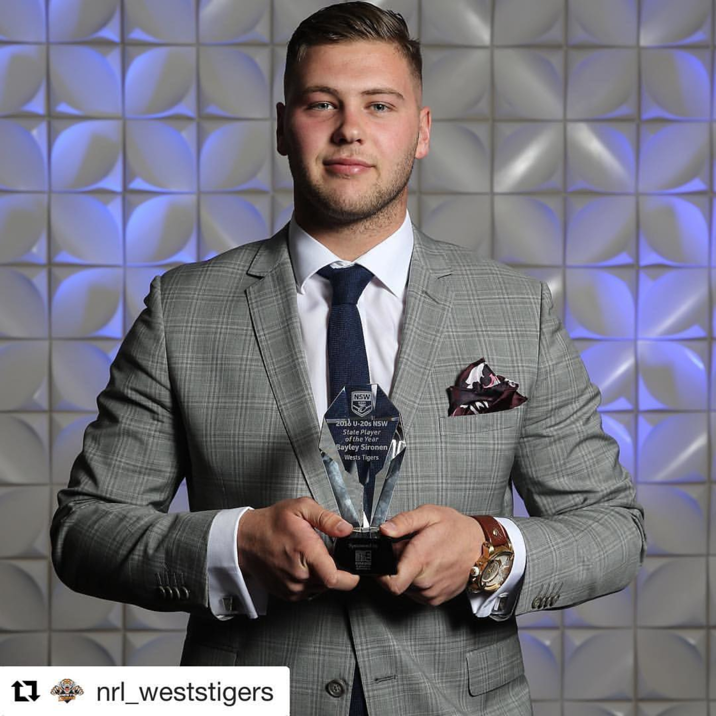 Man in a gray checked suit holding an award against a modern textured backdrop.