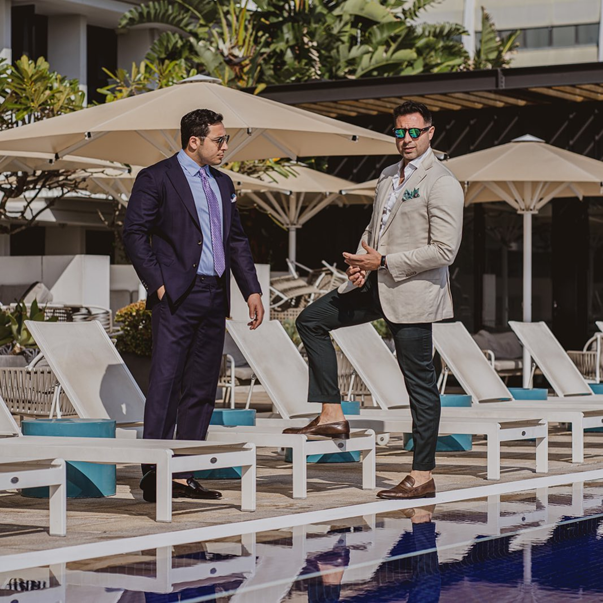 Two men in sharp suits stand by a poolside lounge area at a modern outdoor venue.