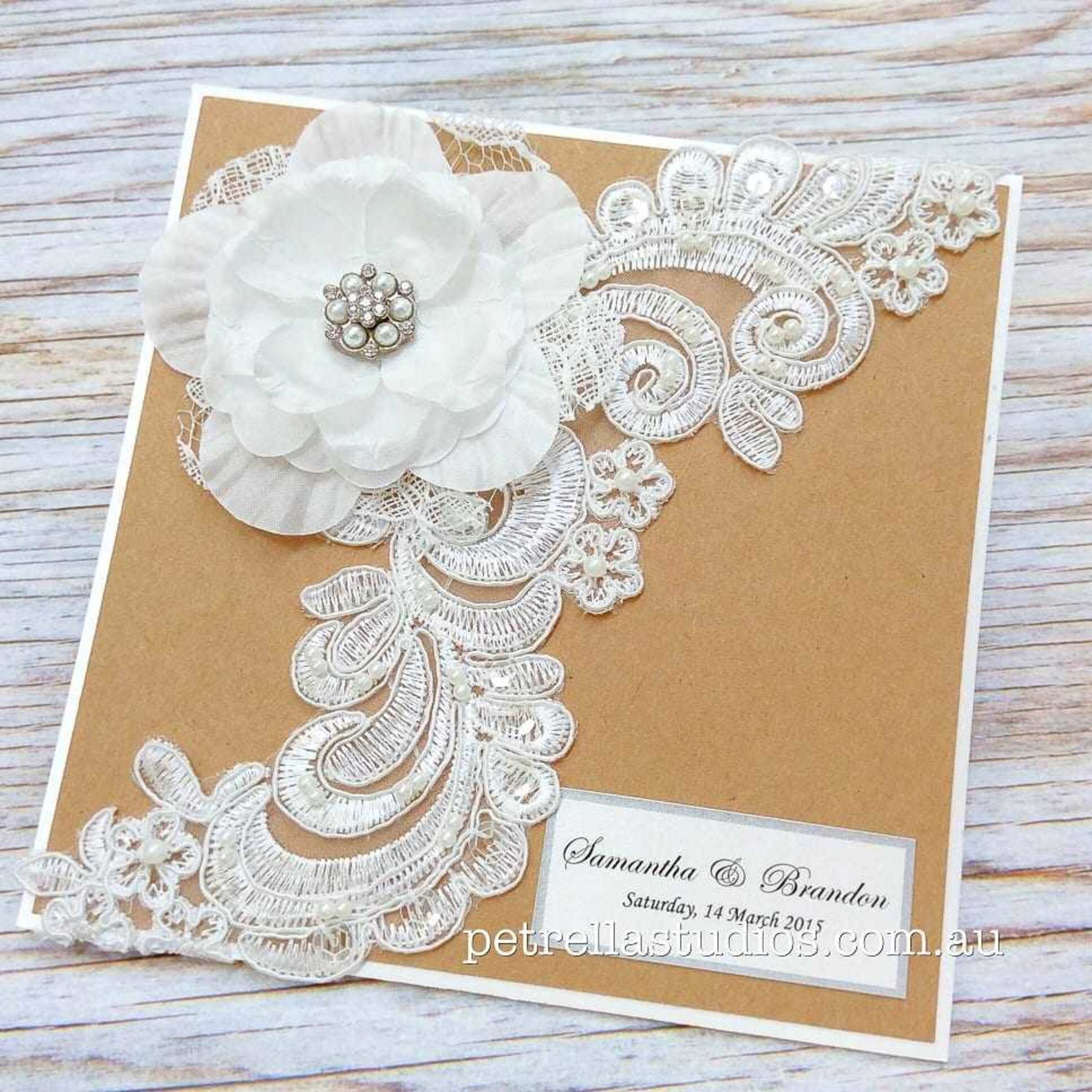 Rustic kraft wedding invitation decorated with white lace applique and a fabric flower with pearl center.