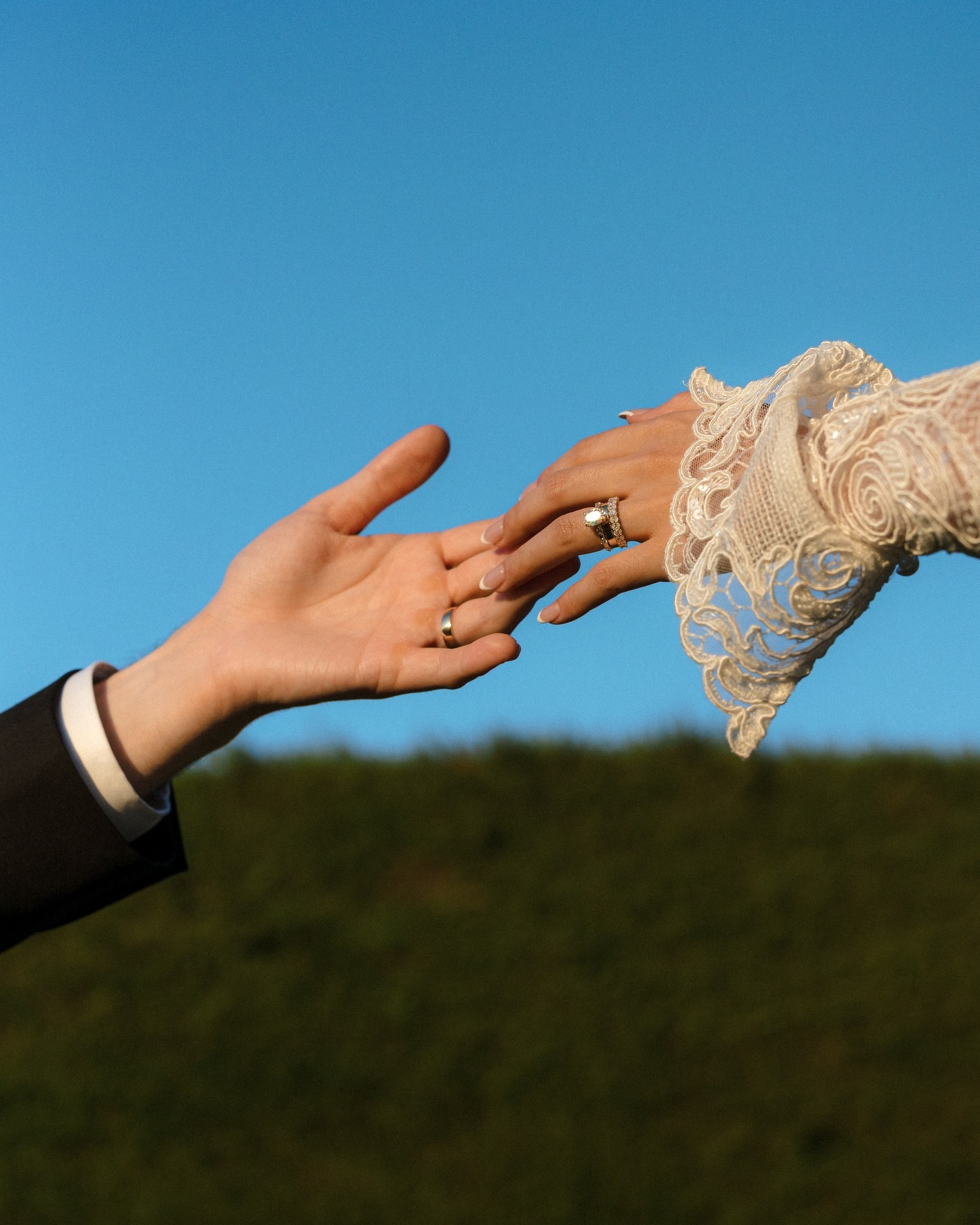 Close-up of a bride and groom reaching for each other’s hands, showing wedding rings against a clear blue sky.