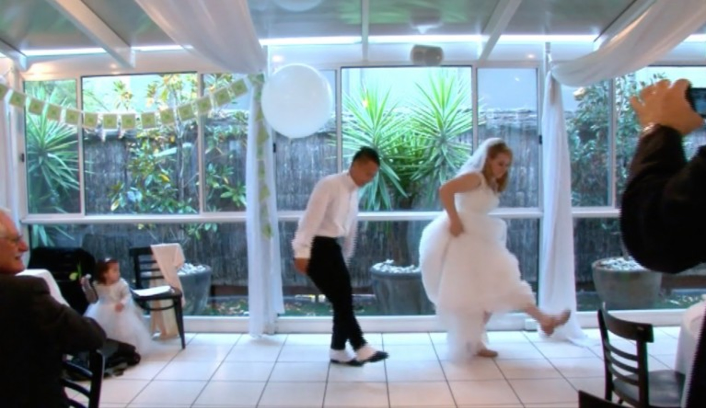 Bride and groom dancing playfully at an indoor wedding reception with guests watching.