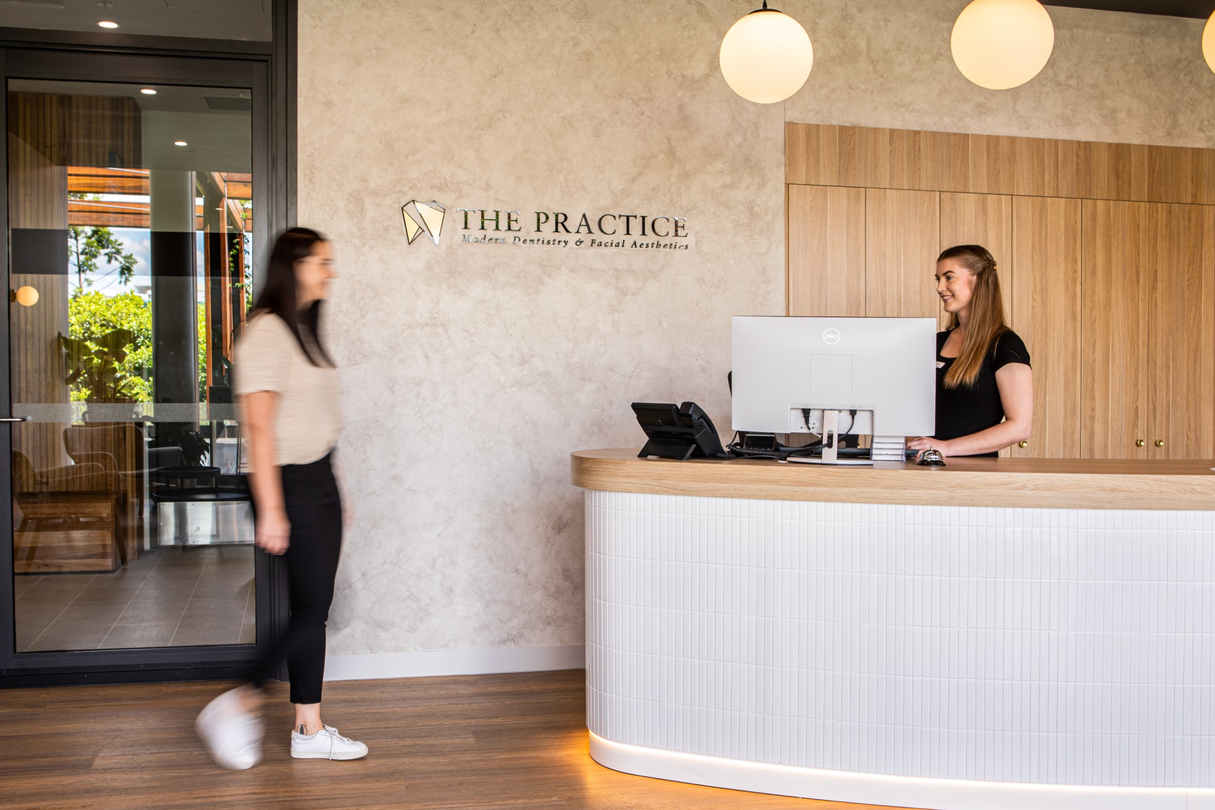 Modern reception area with a staff member greeting a guest at a curved wooden front desk.