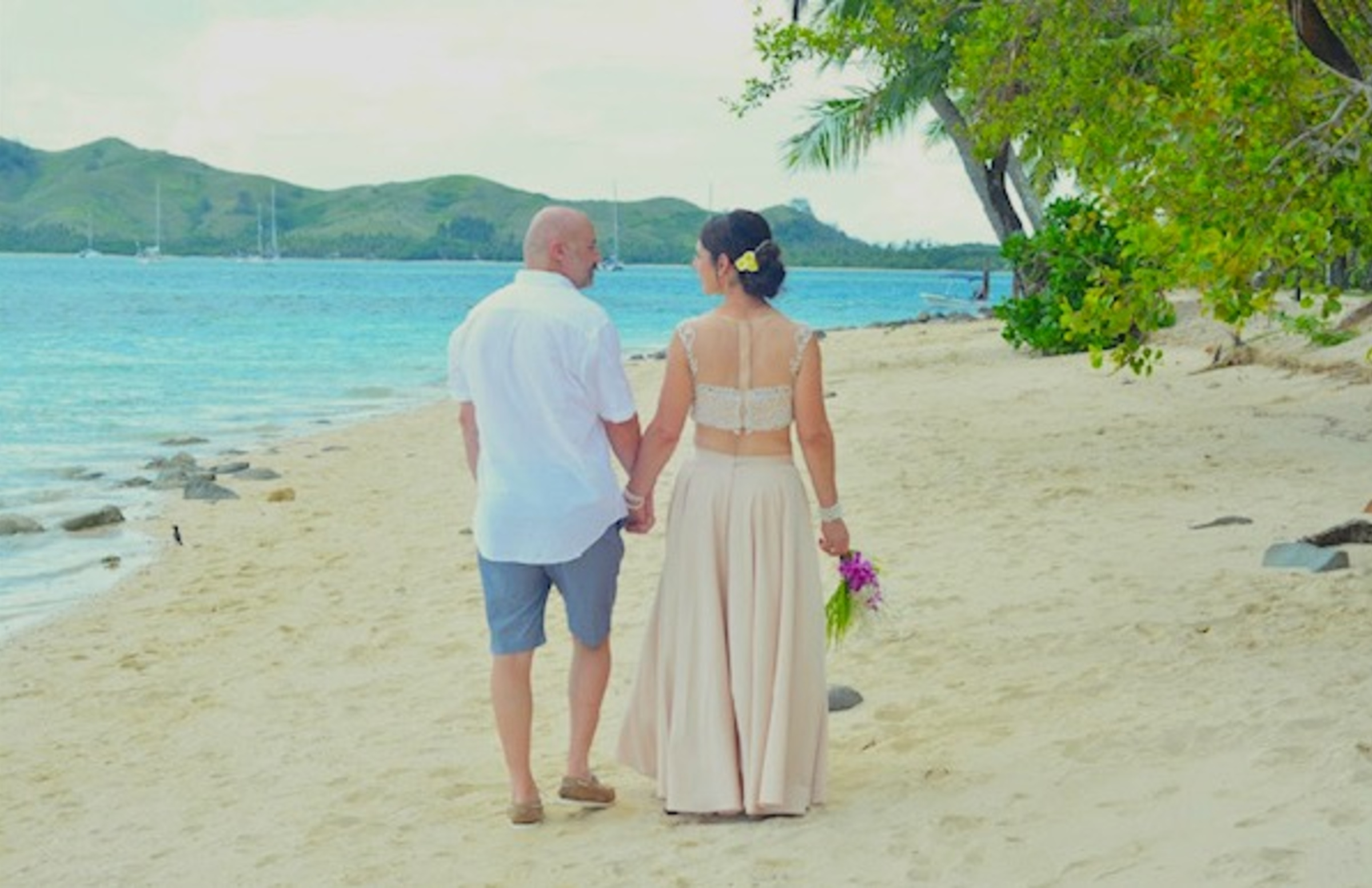 Couple holding hands on a sandy tropical beach after their wedding ceremony by the ocean.