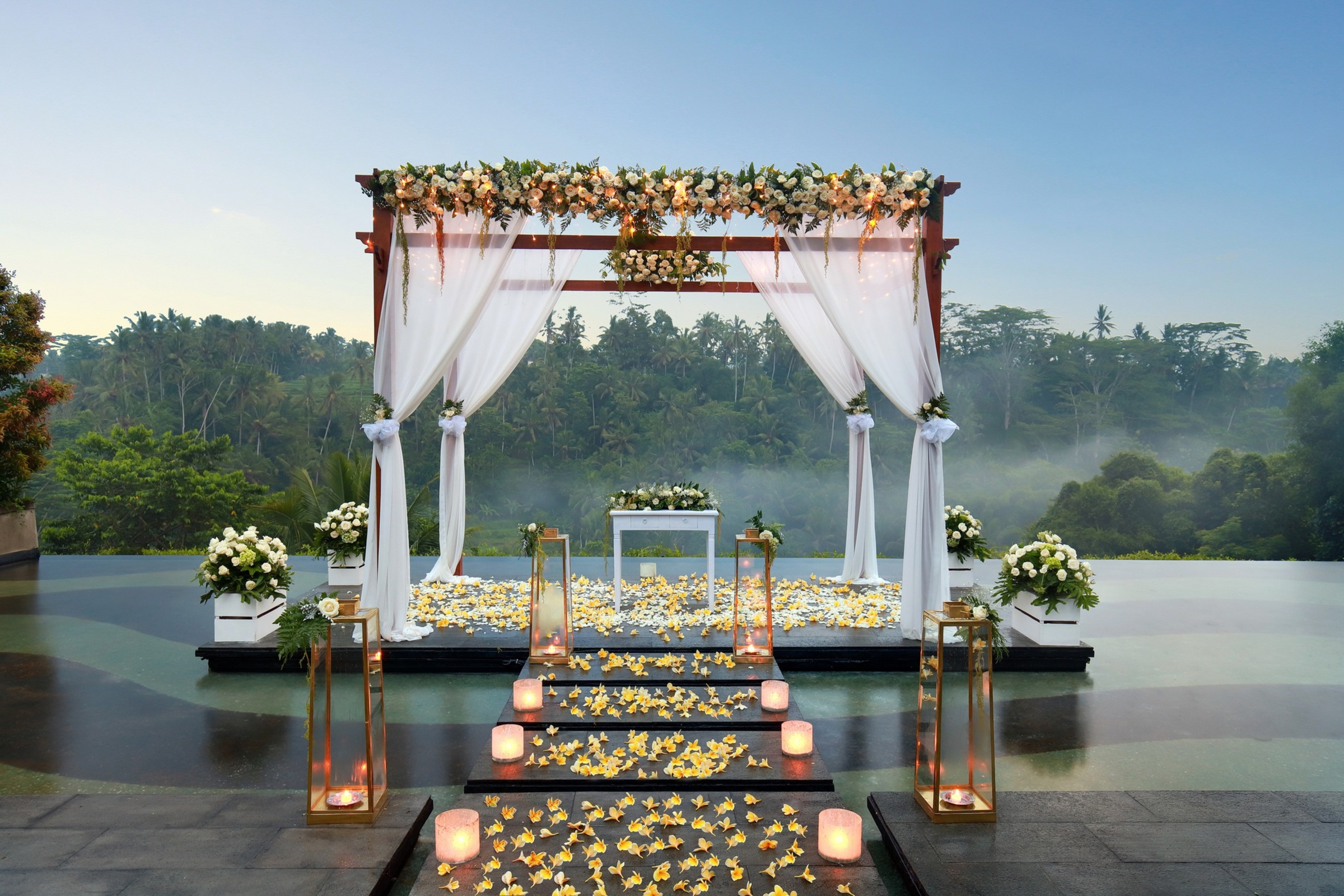Romantic outdoor wedding ceremony arch with white draping, florals, and candlelit aisle over a tropical infinity pool.