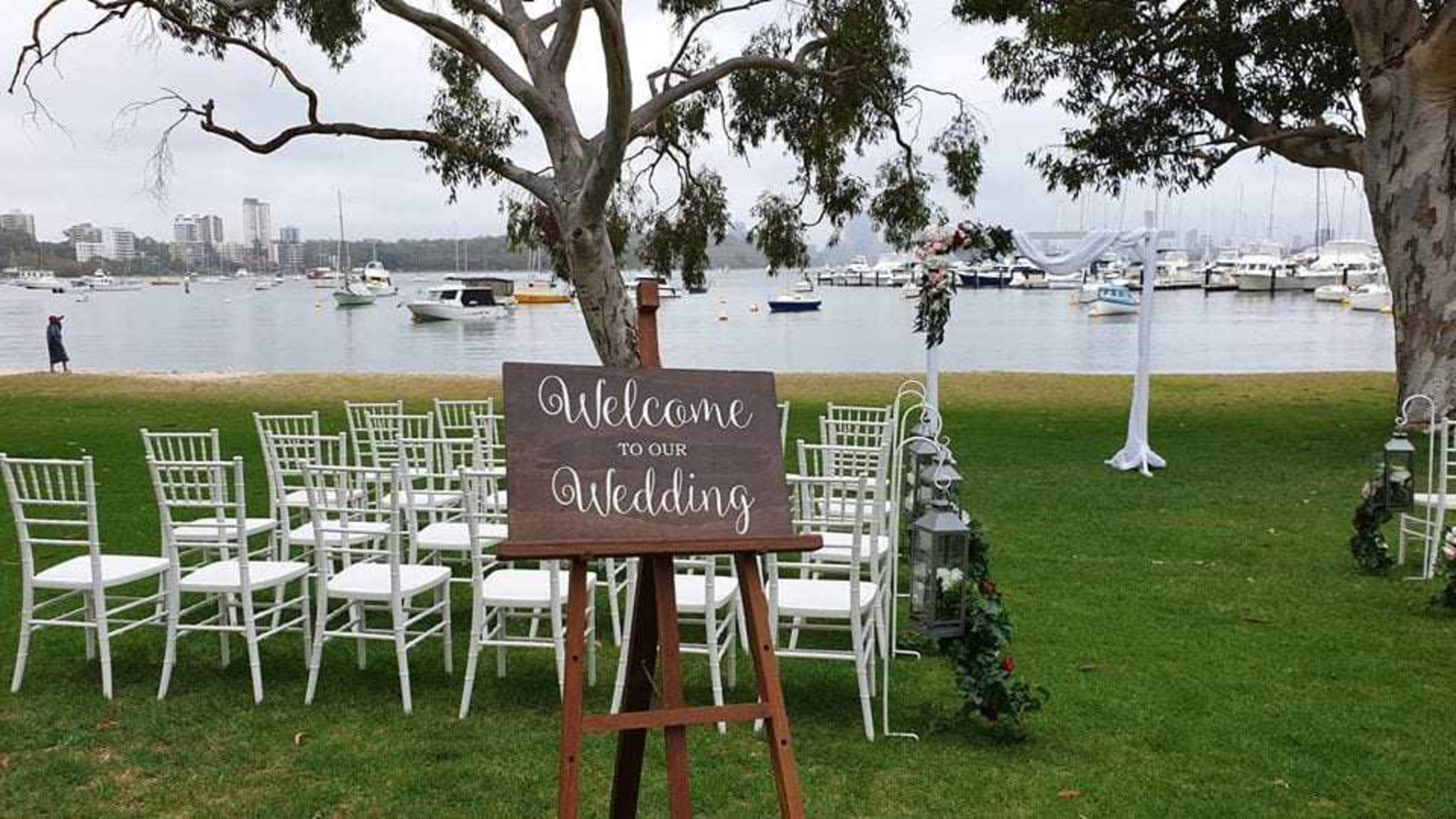 Outdoor waterfront wedding ceremony setup with white chairs, welcome sign, and arch by the harbor.