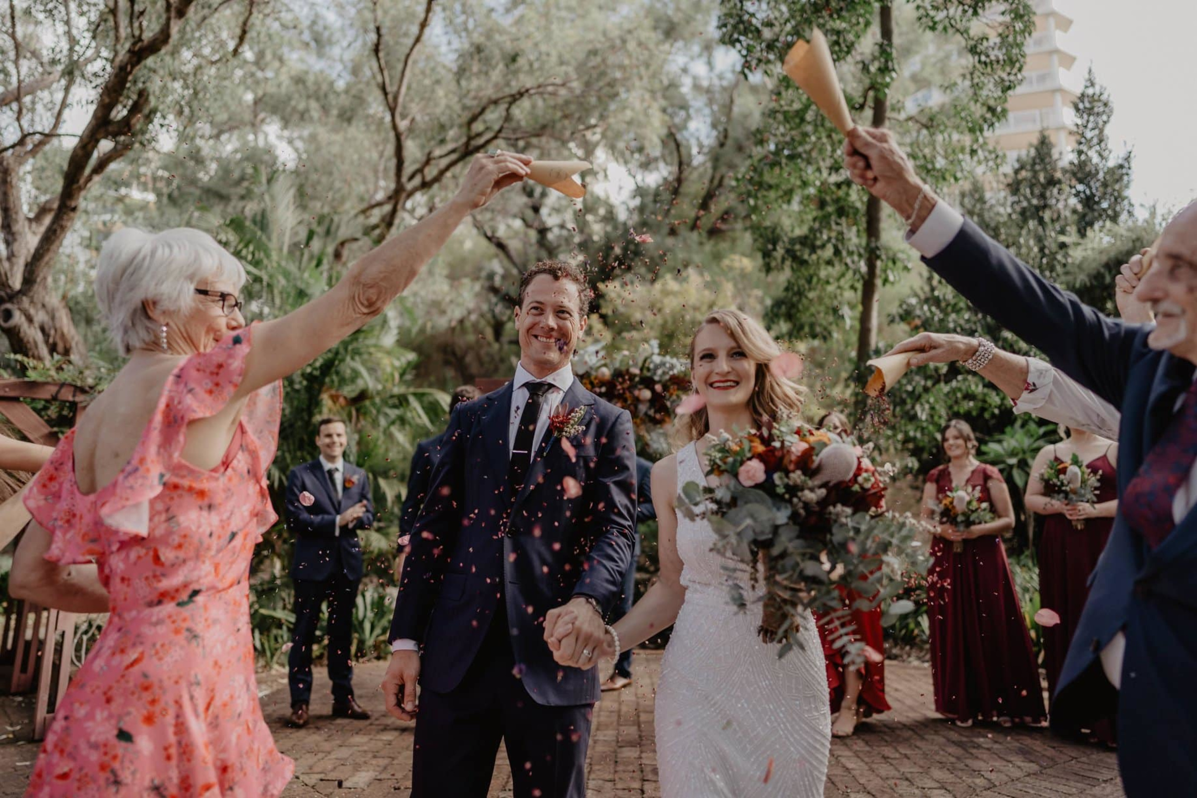 Newlywed couple walks down an outdoor aisle smiling as guests toss flower petal confetti around them.