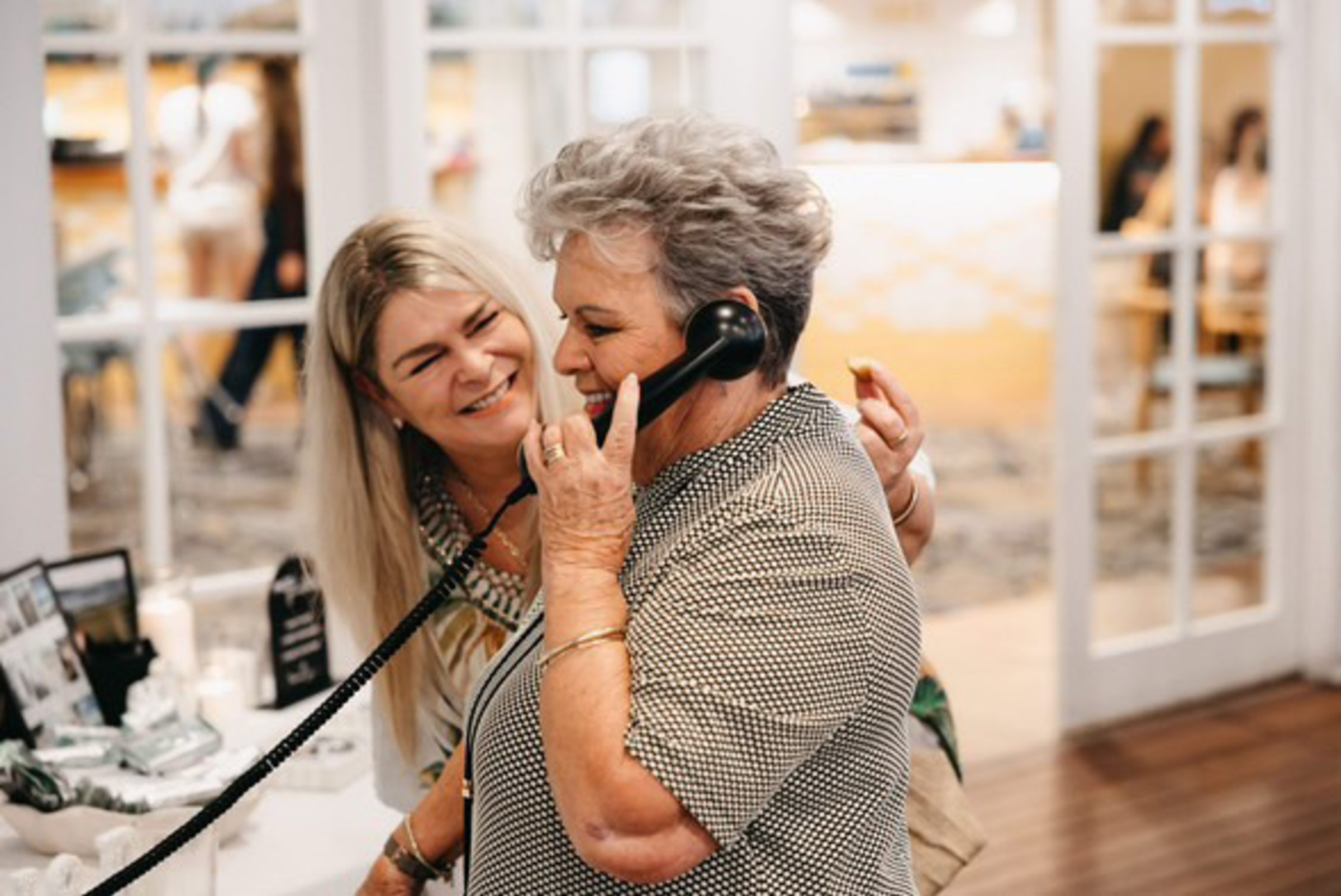 Two women smile as one records a message on a vintage-style phone at a wedding reception.