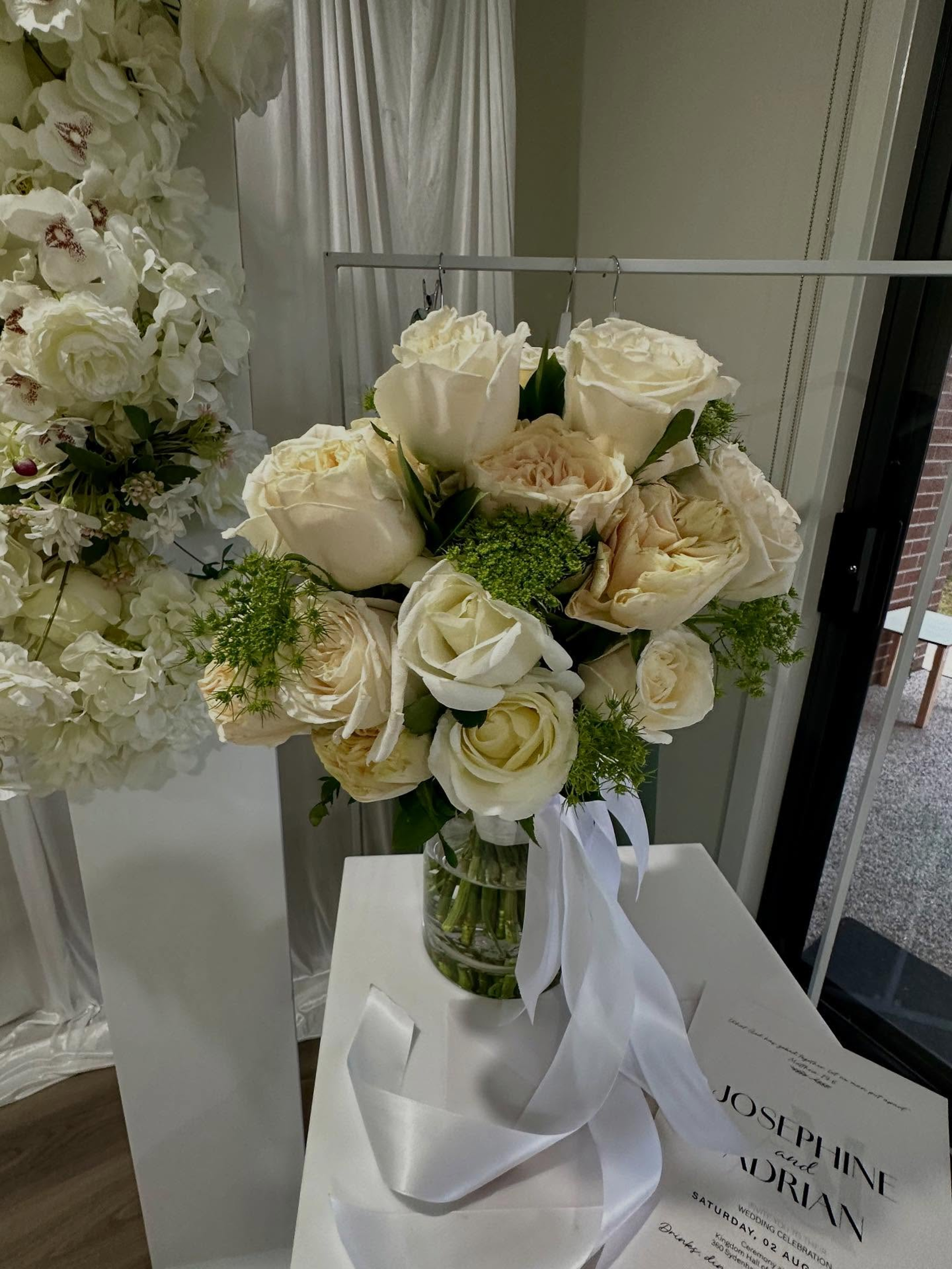 Elegant white and cream rose bridal bouquet with flowing white ribbons displayed beside a wedding invitation.