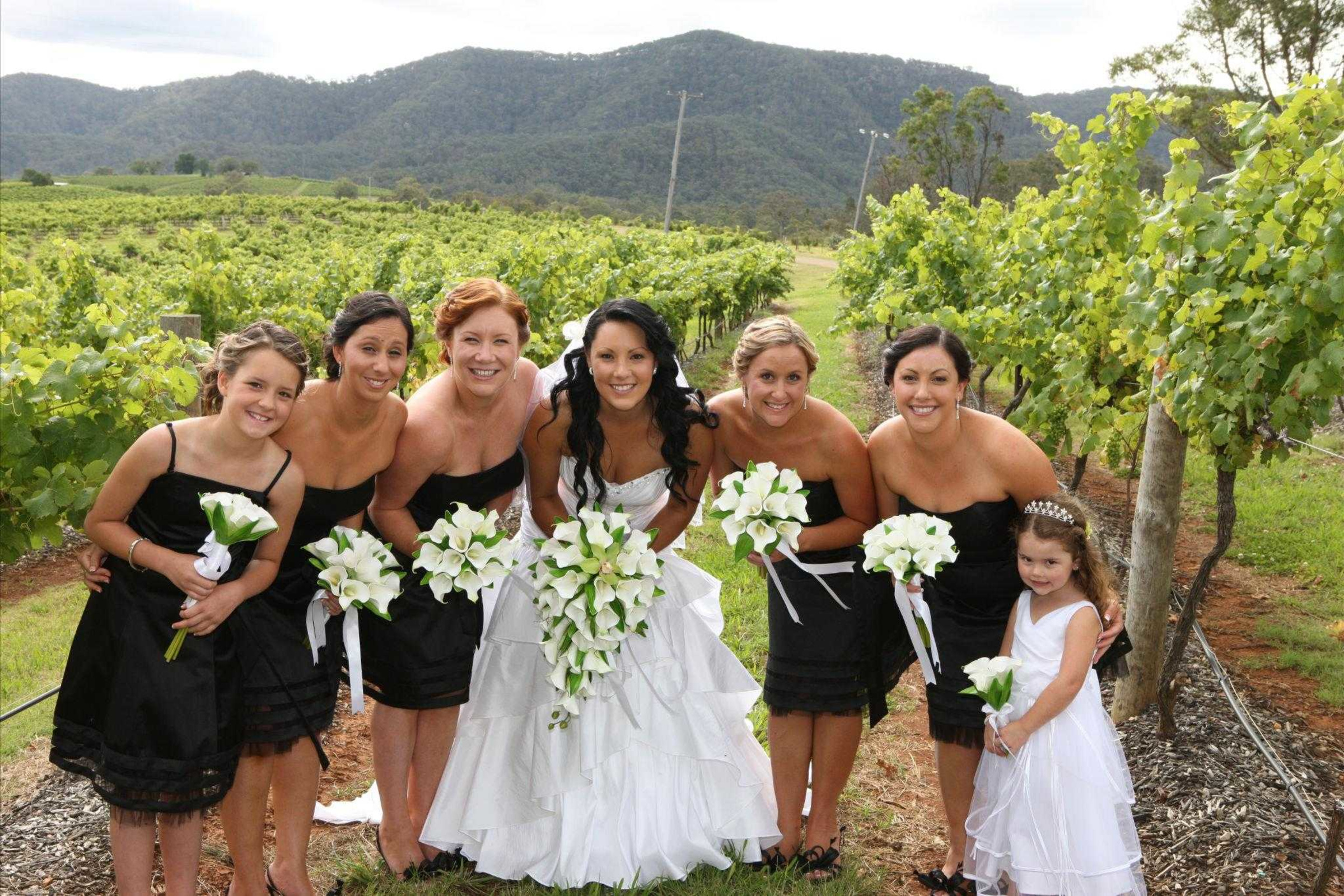 Bride and bridesmaids in black dresses pose with white bouquets in a green vineyard with mountains in the background.