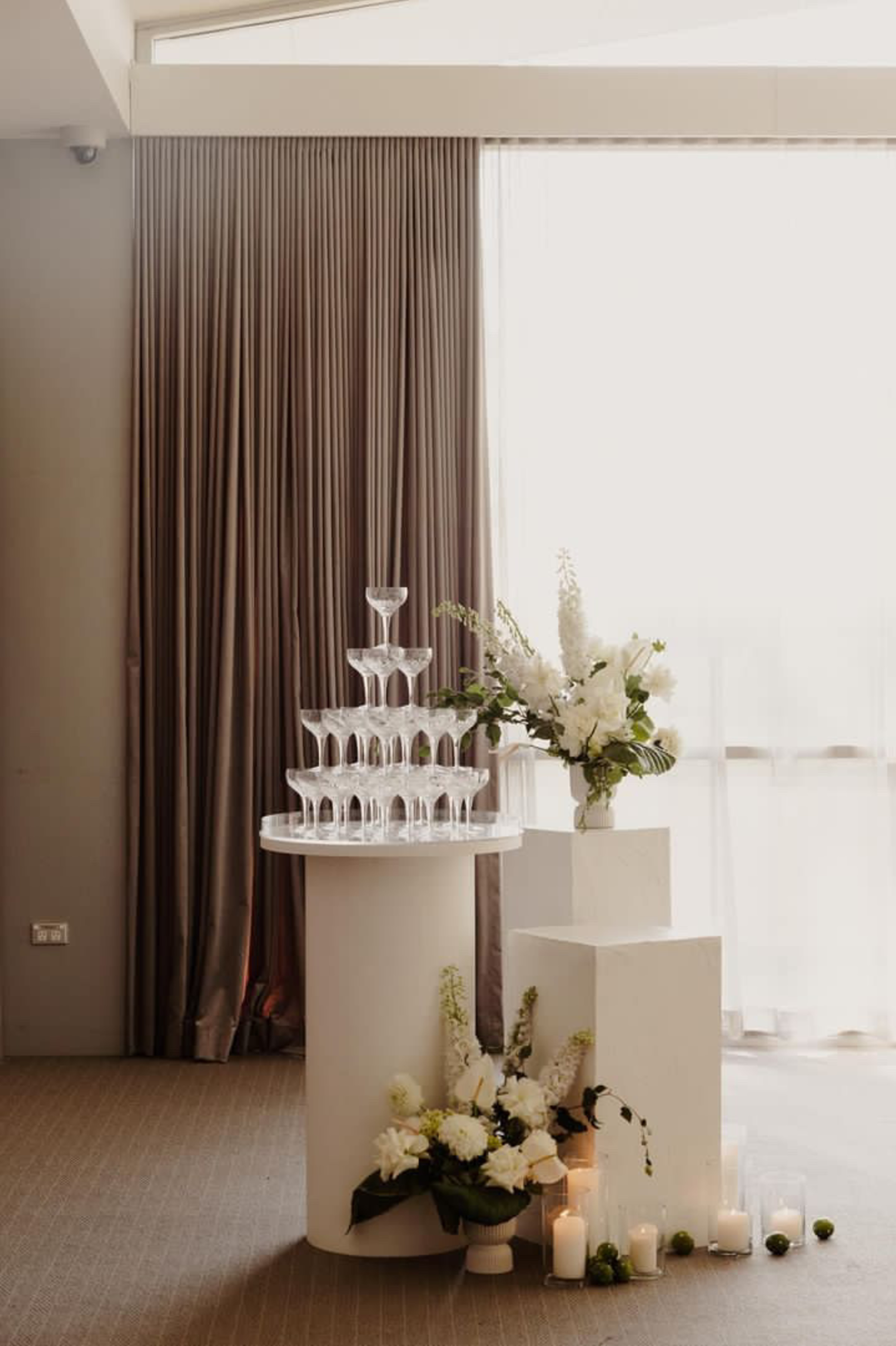 Elegant champagne tower with white floral arrangements and candles in a softly lit indoor wedding setting.