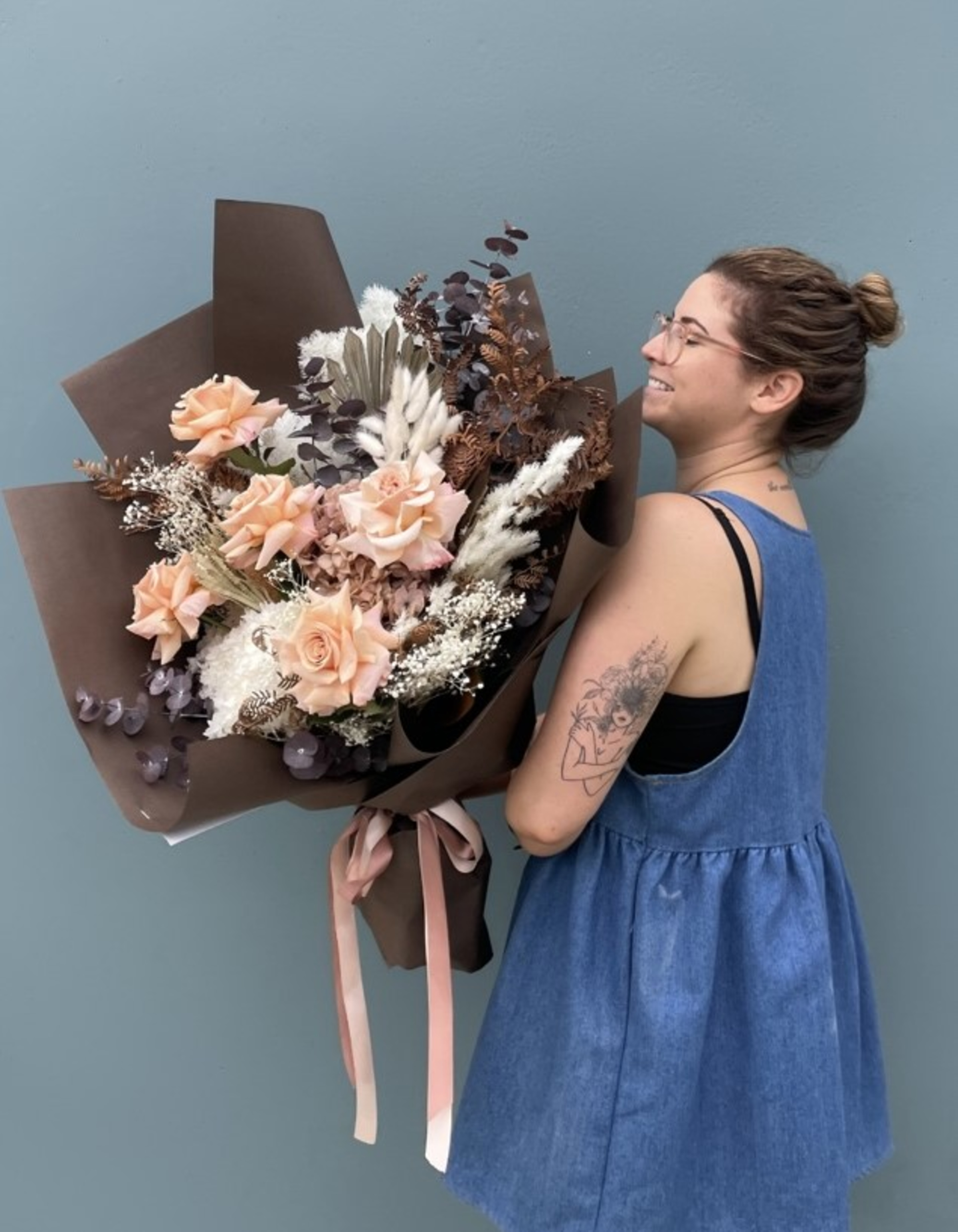 Florist holding a large bouquet of peach roses and dried foliage wrapped in brown paper with pink ribbons.