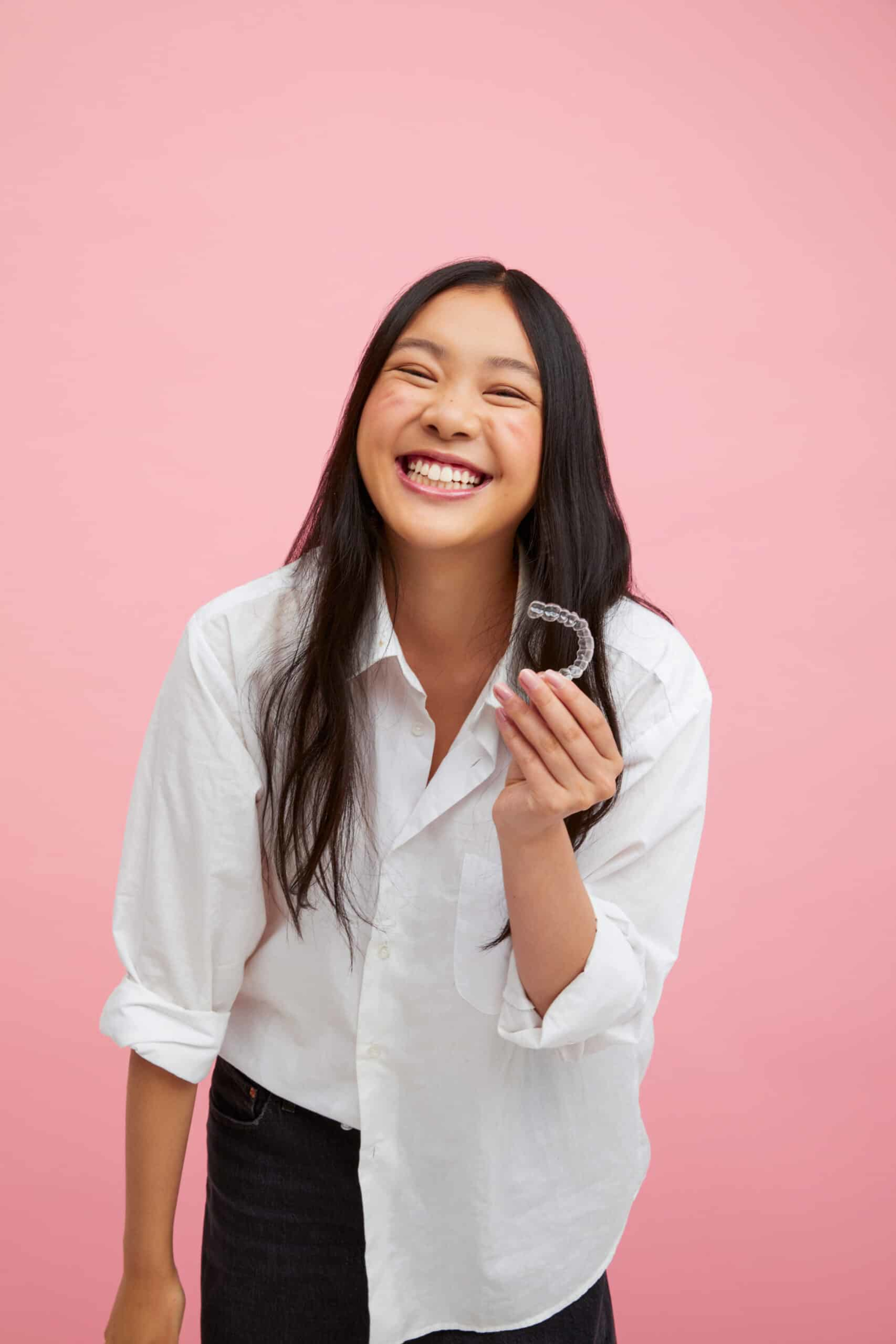 Smiling woman in a white shirt holding a clear dental aligner against a pink background.