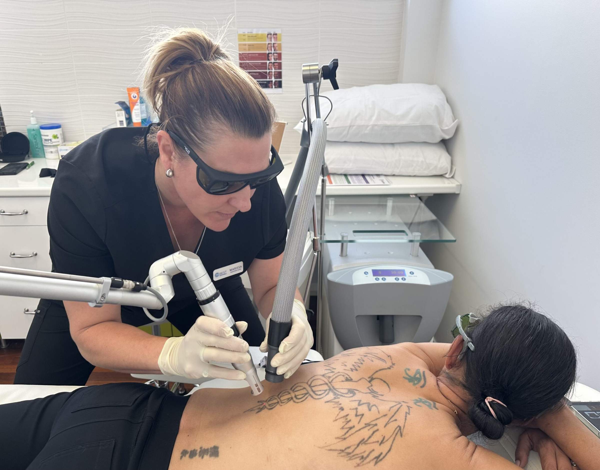 Technician performs laser tattoo removal on a patient lying face down in a cosmetic treatment room.