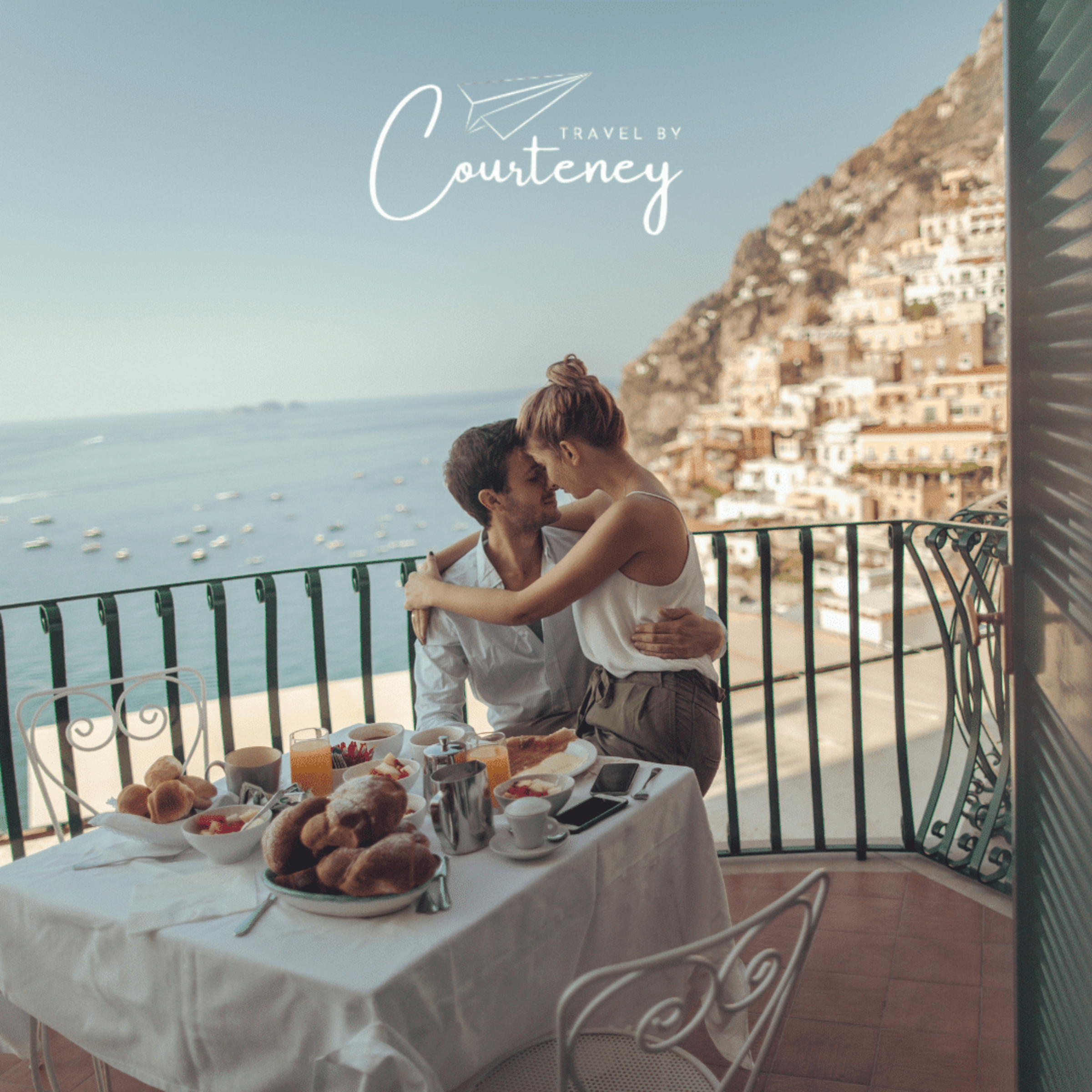 Couple sharing a romantic breakfast on a seaside balcony overlooking a cliffside town and boats.