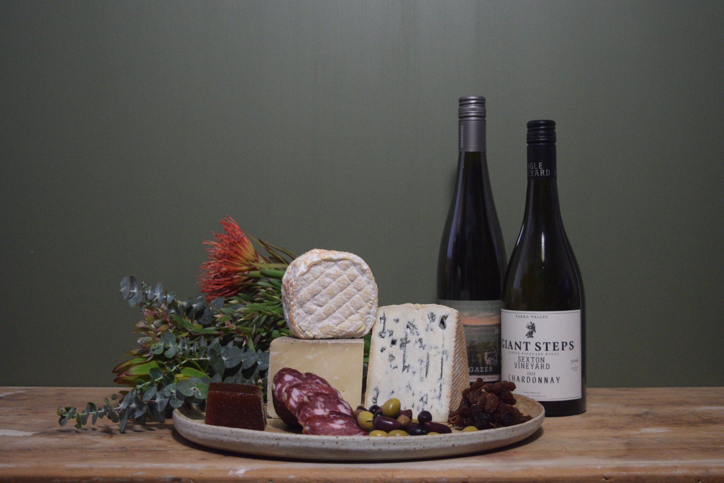 Rustic cheese and charcuterie board with wine bottles, olives, and flowers on a wooden table.