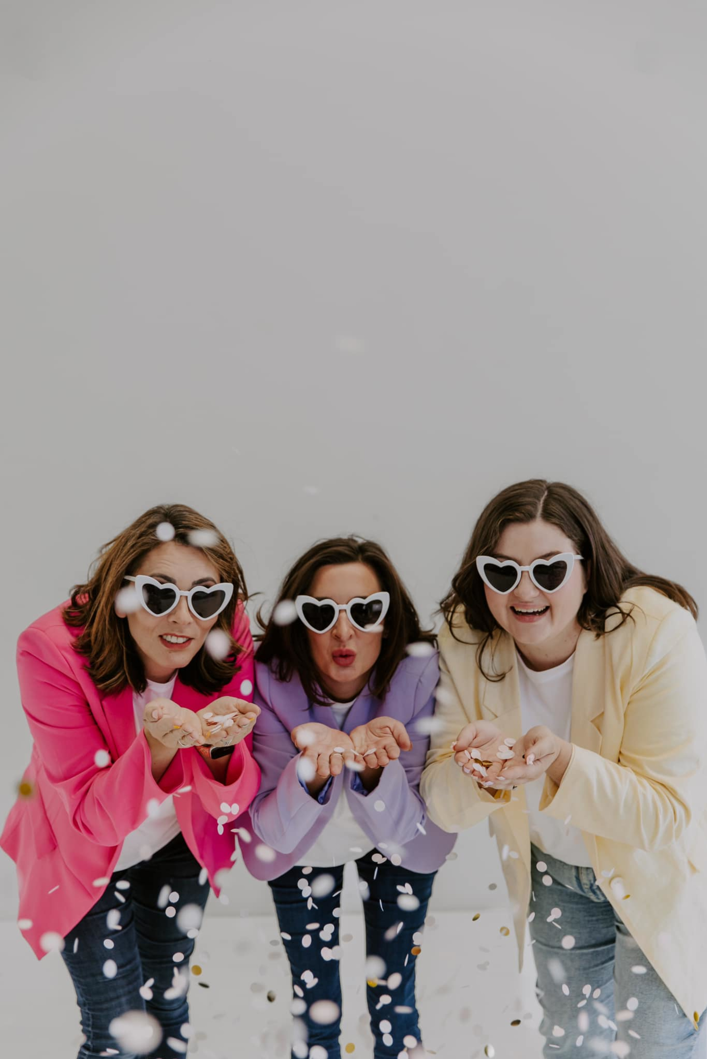 Three women in colorful blazers and heart sunglasses blow confetti toward the camera in a playful studio shot.