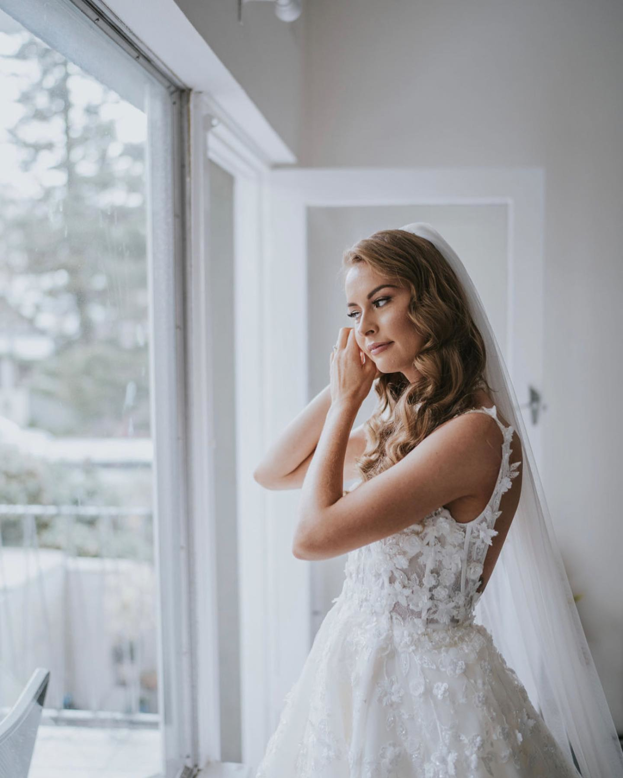 Bride in a lace wedding dress adjusts her earring while standing by a bright window indoors.
