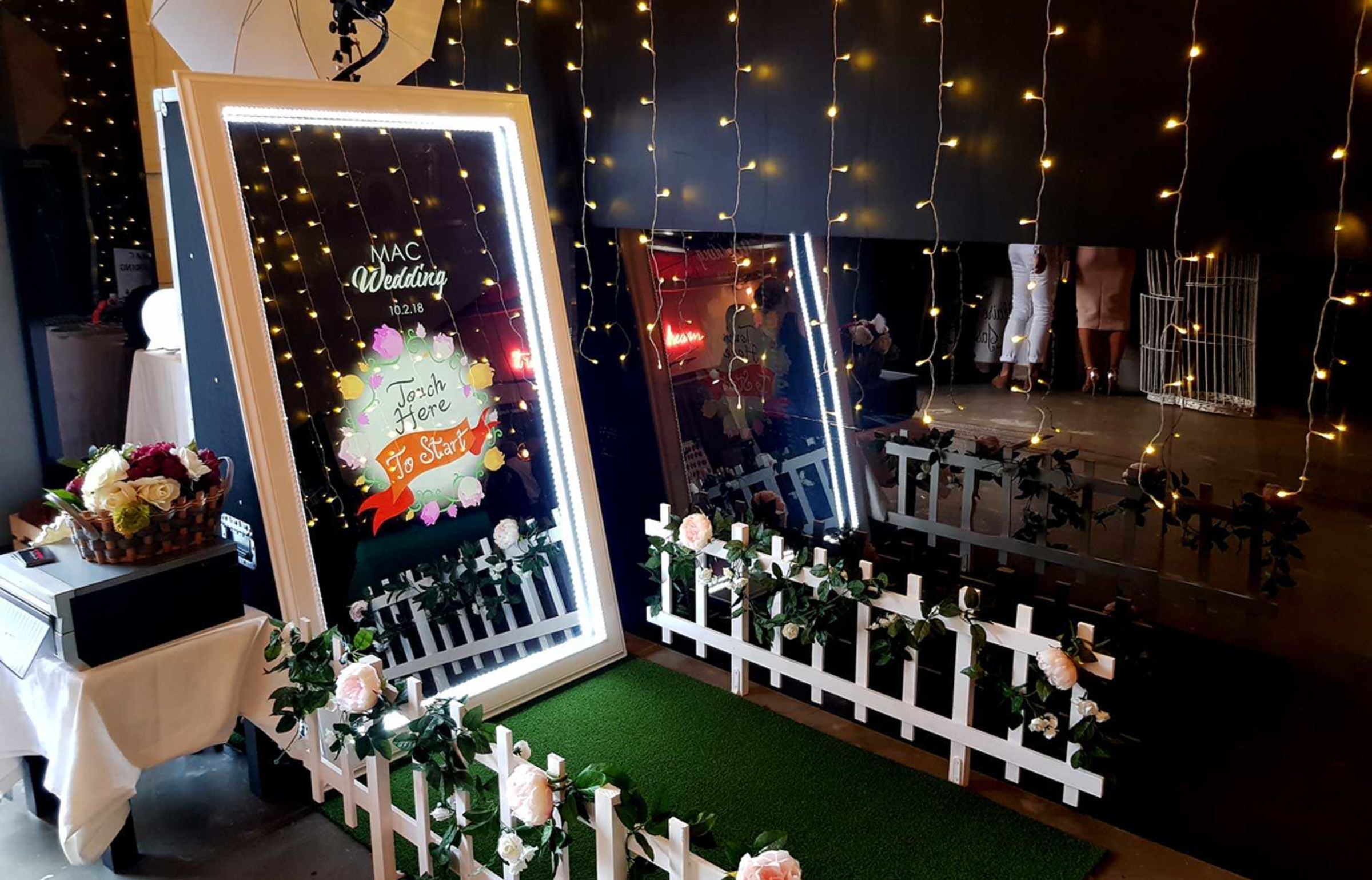 Interactive mirror wedding photo booth framed with fairy lights, white picket fence, and floral decorations.