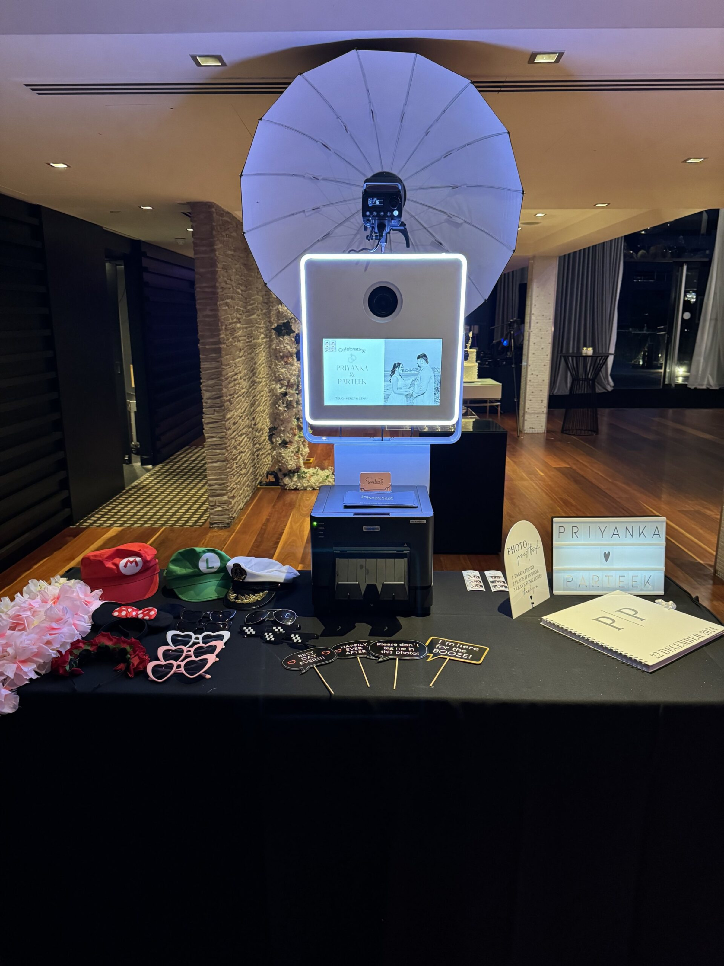 Wedding reception photobooth setup with props table, printer, and illuminated sign for guests.