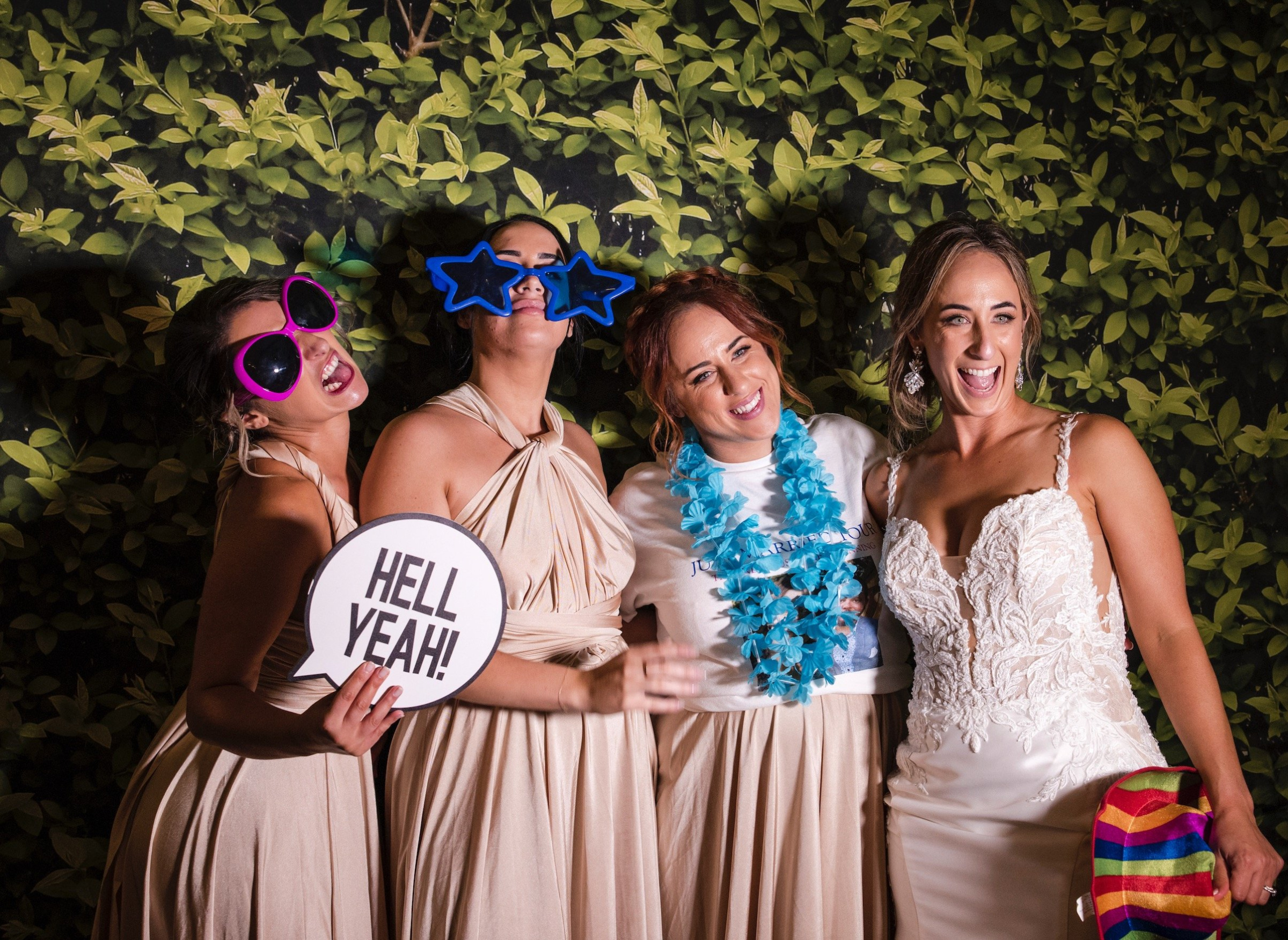 Bride and bridesmaids pose with fun photo booth props in front of a leafy green backdrop.