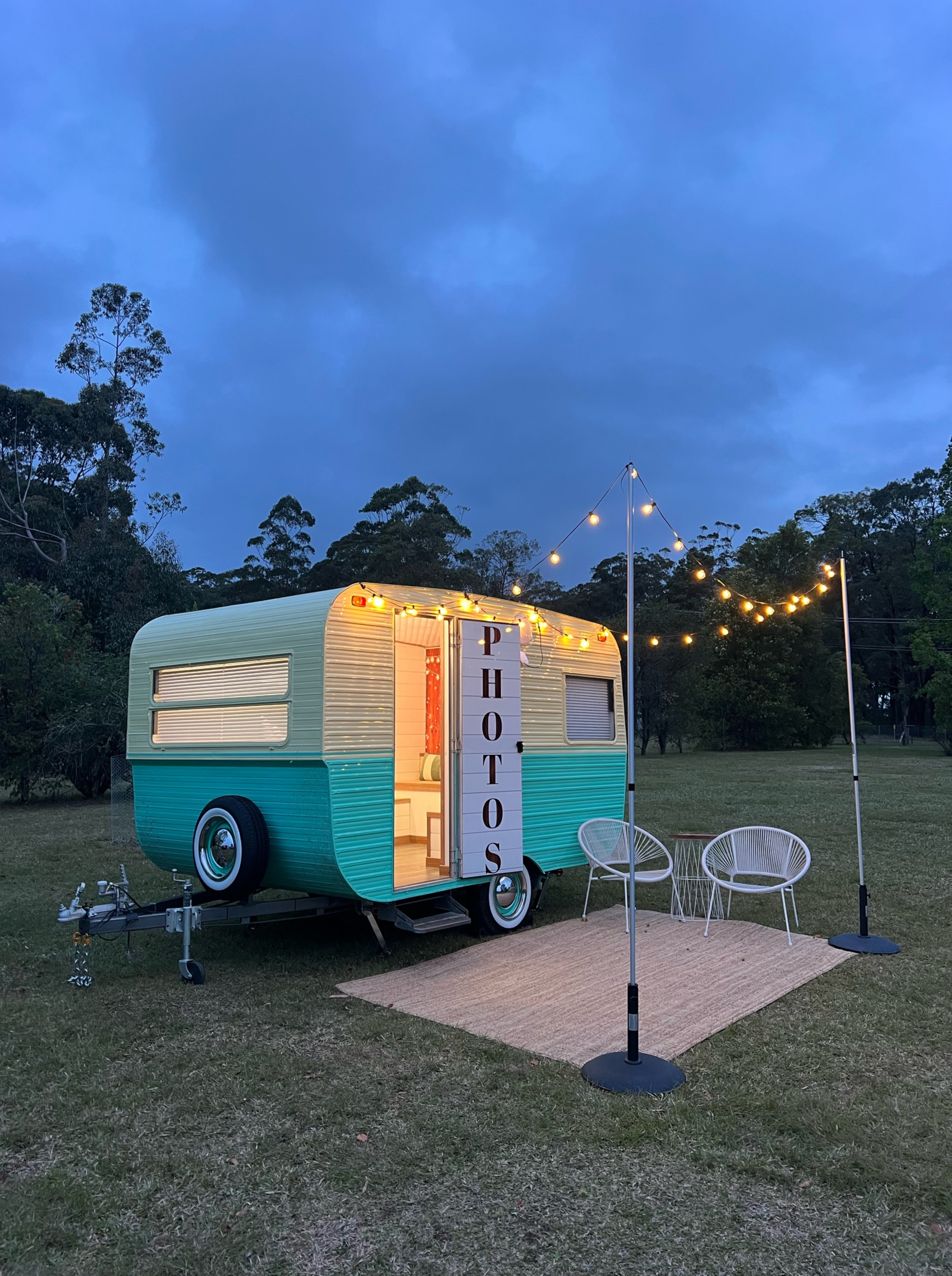 Vintage caravan photo booth with string lights and chairs set up on a lawn at dusk.