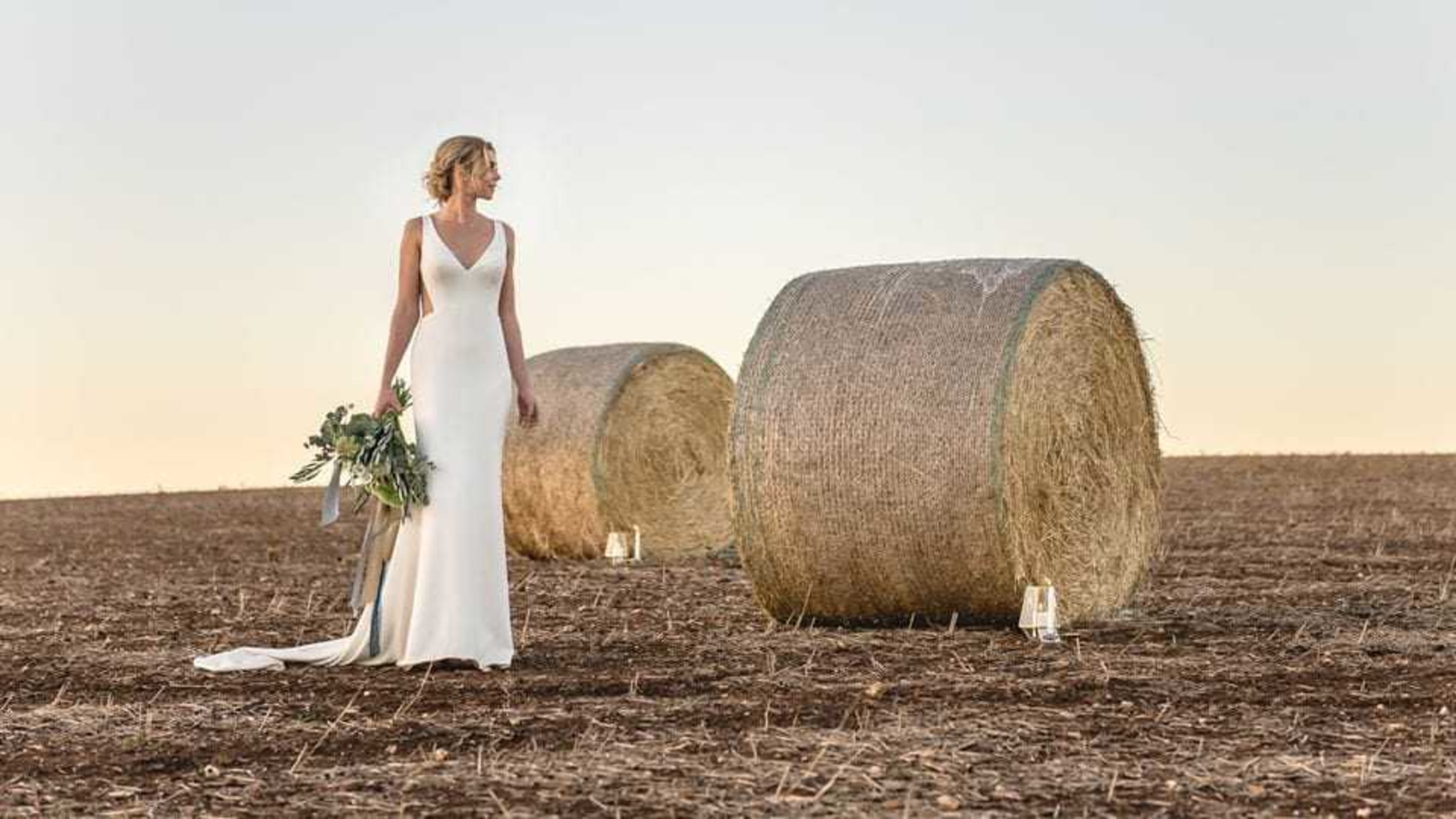 Bride in a simple white gown holding a bouquet while standing in a field with large hay bales at sunset.