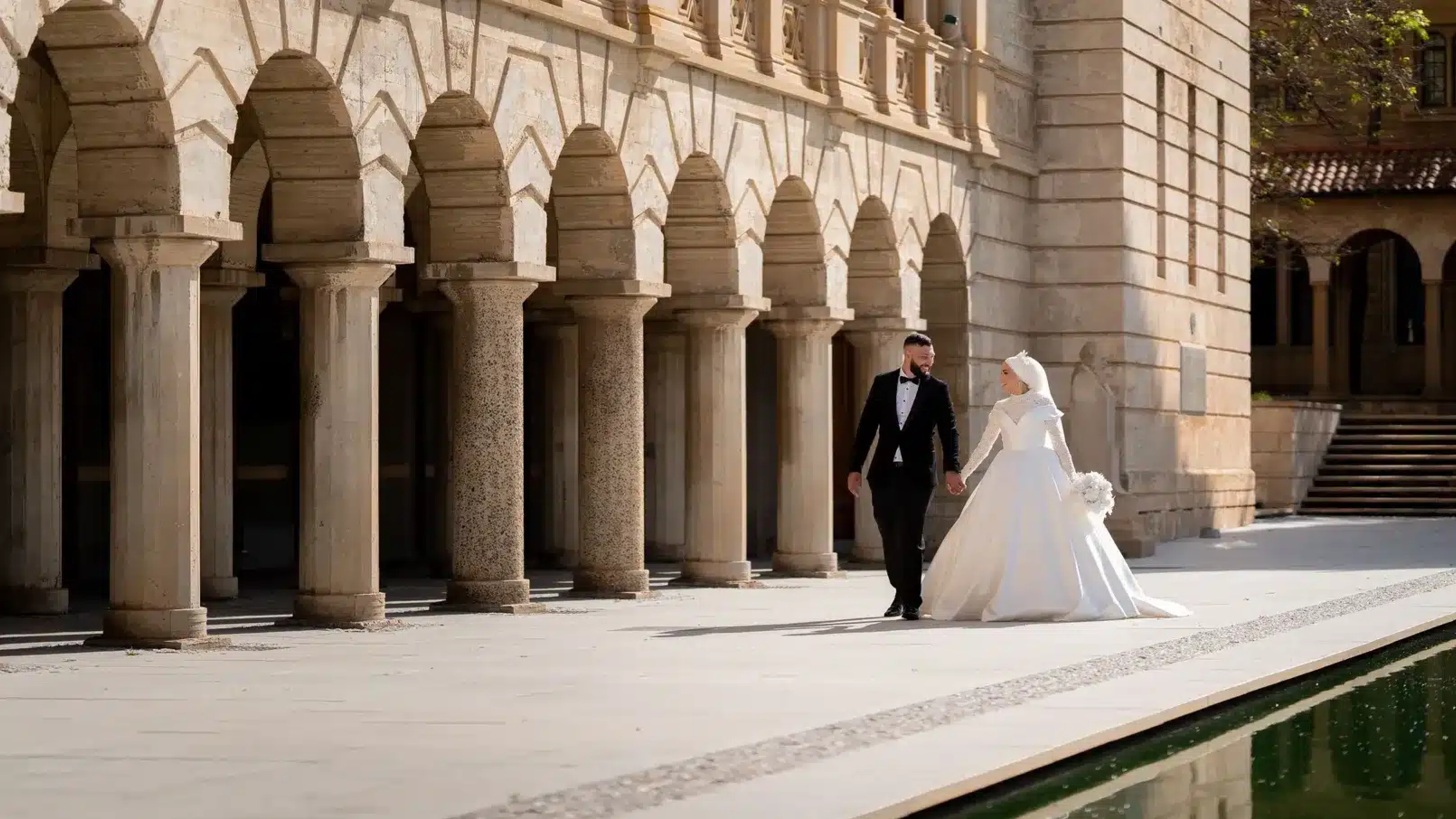 Bride and groom walk hand in hand beside a grand stone colonnade and reflective pool at an elegant wedding venue.