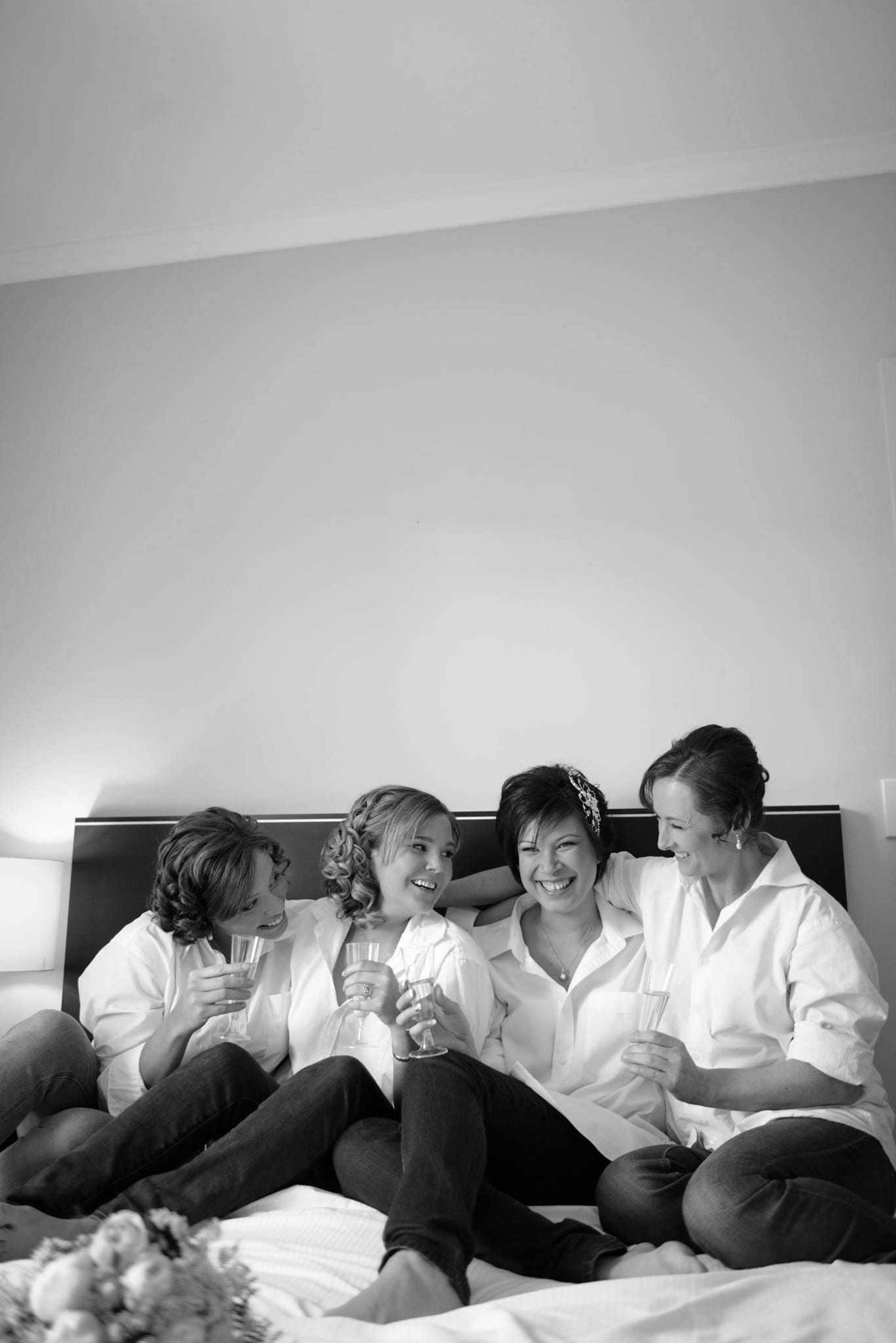 Candid black and white photo of a bride and bridesmaids laughing on a bed with champagne glasses.