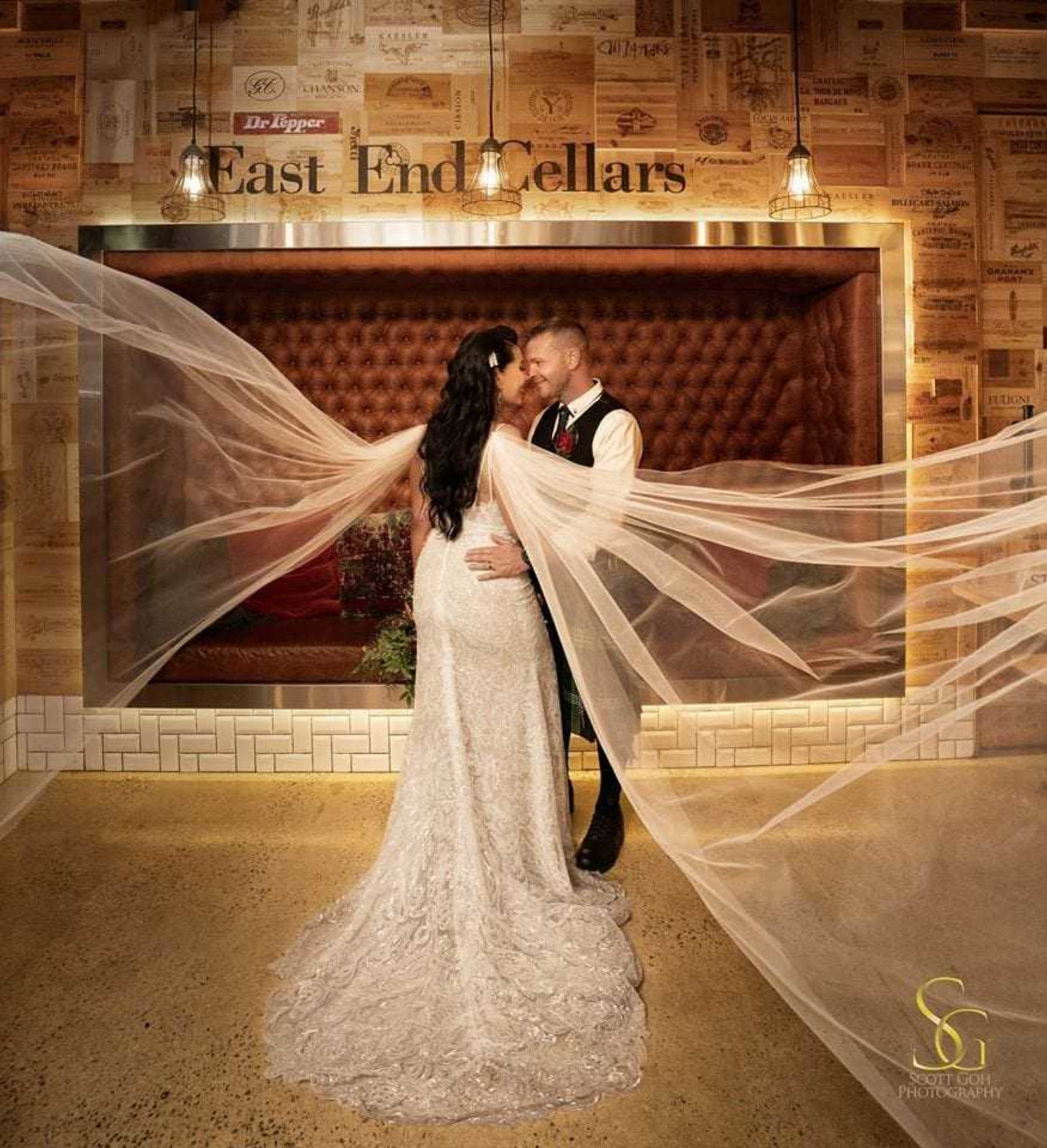 Bride and groom embrace at an indoor winery venue as the bride’s long veil billows dramatically around them.