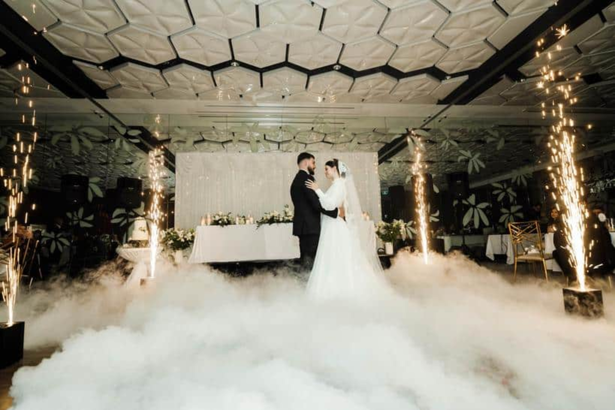 Bride and groom share their first dance amid fog and indoor sparklers in a modern wedding ballroom.