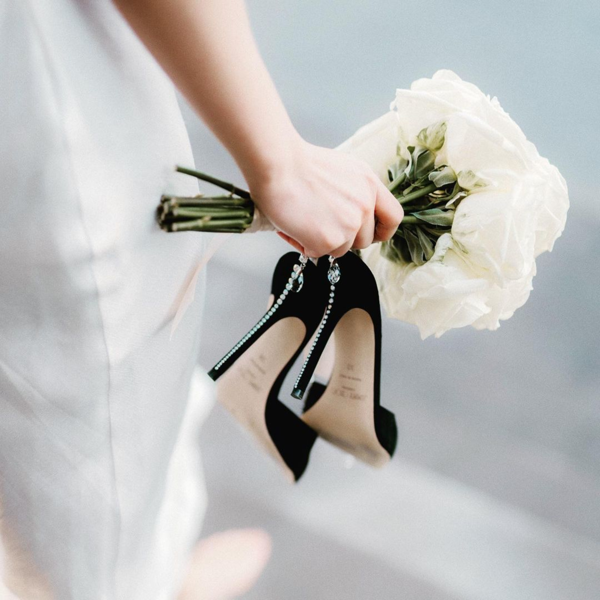 Bride holding white bouquet and black high heels with crystal straps in a soft, modern wedding detail shot.